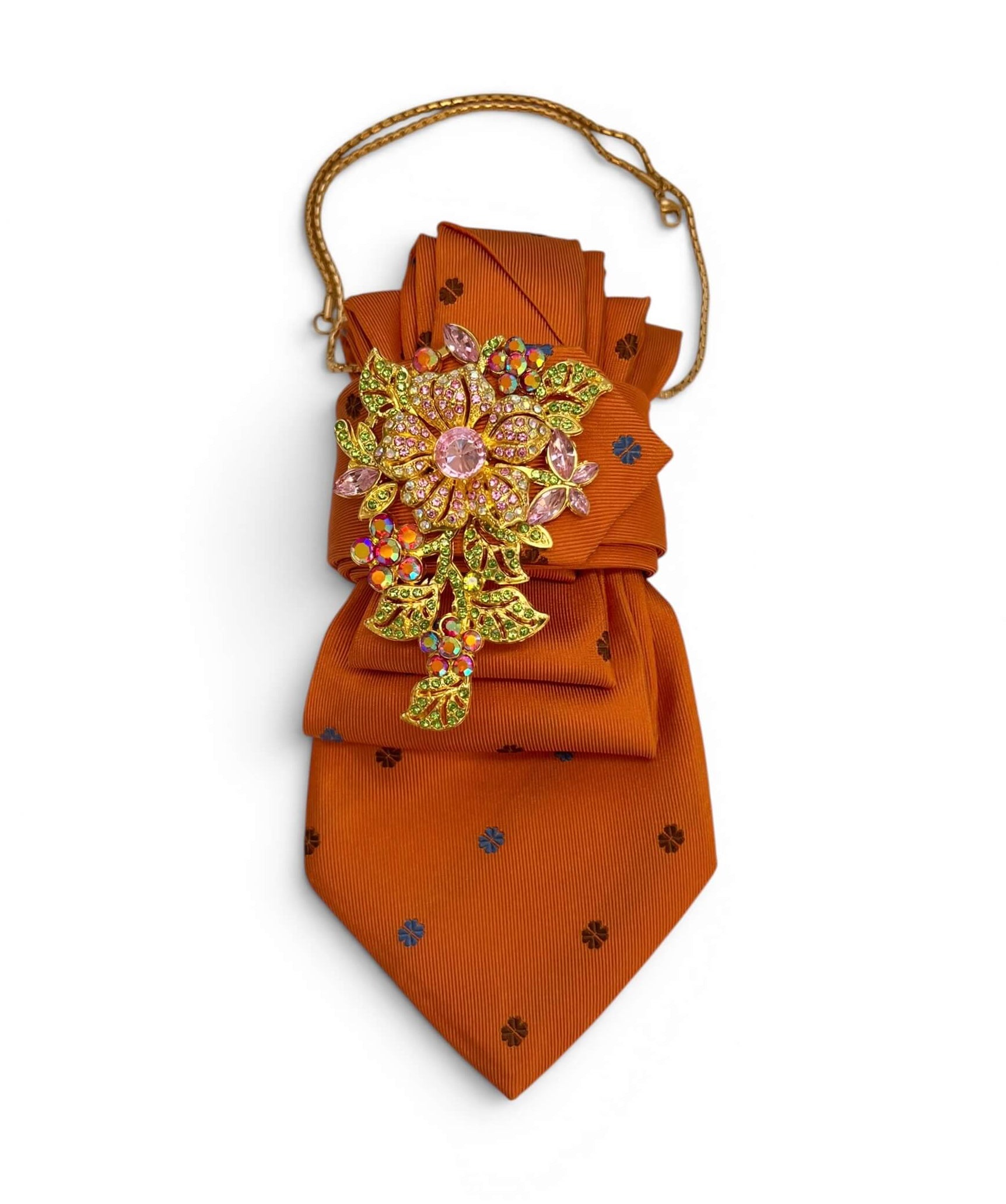 Orangefarbene Krawatte mit goldfarbener Kette und Brosche mit rosa, goldfarbenen und schillernden Schmucksteinen auf hellem Hintergrund.