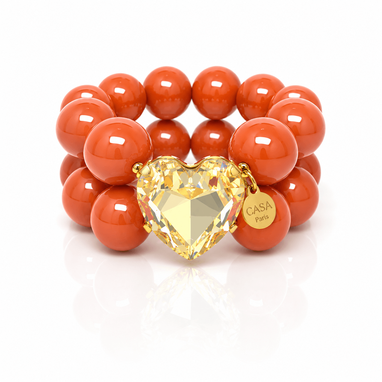Armband aus glänzenden orangefarbenen Resin-Perlen mit großem facettiertem Kristallherz in Champagner-Optik, goldfarbener Fassung und rundem Casapari-Anhänger auf weißem Hintergrund.