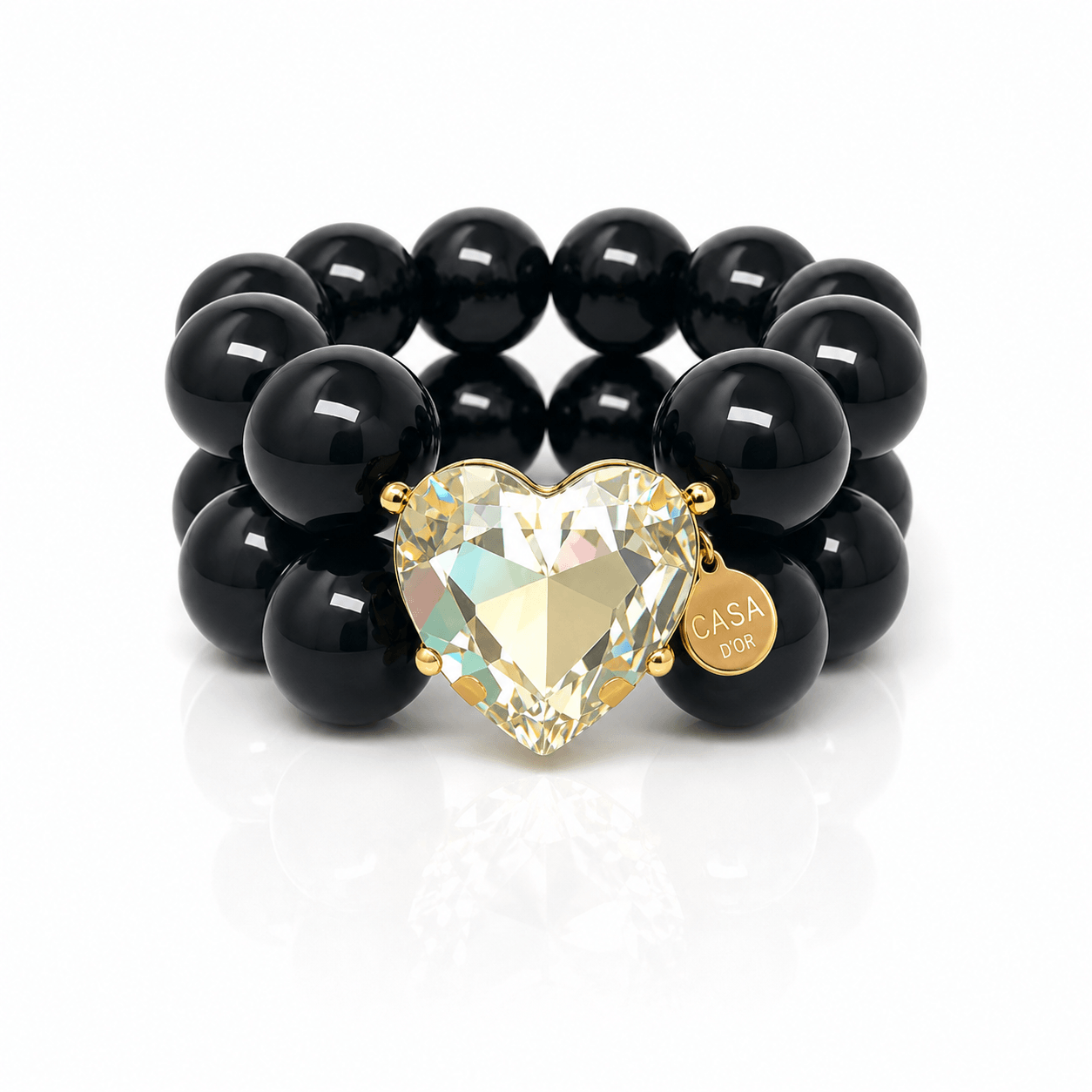 Armband aus glänzenden schwarzen Resin-Perlen mit großem facettiertem Kristallherz, goldfarbener Fassung und rundem Casapari-Anhänger auf weißem Hintergrund.