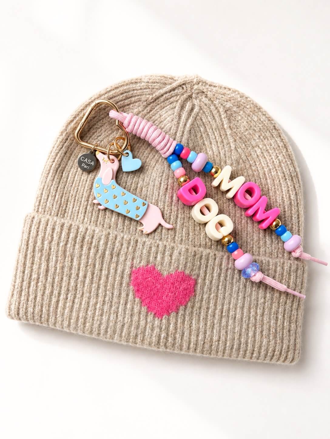 Beige Strick-Beanie mit pinkfarbenem Herz, dekoriert mit farbigem CASAPARI Taschenanhänger und Buchstaben-Charms.
