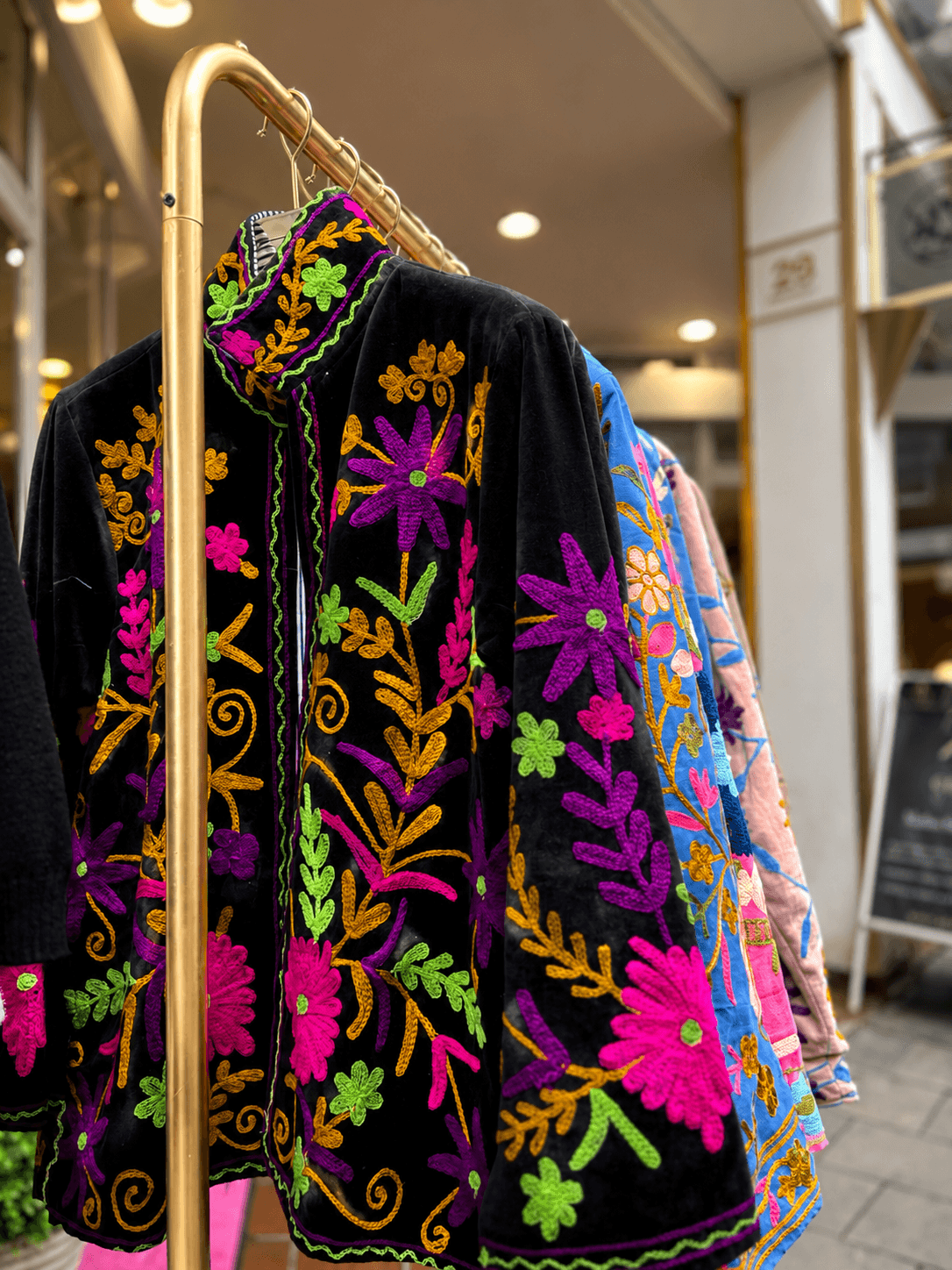 Schwarze CASAPARI Jacke „Black Garten“ mit farbenprächtiger floraler Stickerei in Pink, Violett, Grün und Gold auf einem goldfarbenen Kleiderständer vor der Boutique.