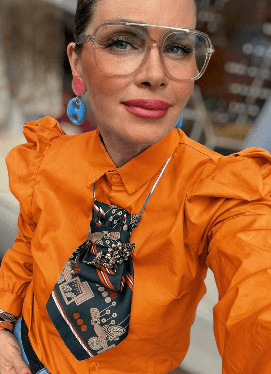 Frau trägt eine leuchtend orange Bluse mit Puffärmeln, kombiniert mit heller Hose und Accessoires, aufgenommen in einem hellen Raum mit großen Fenstern.