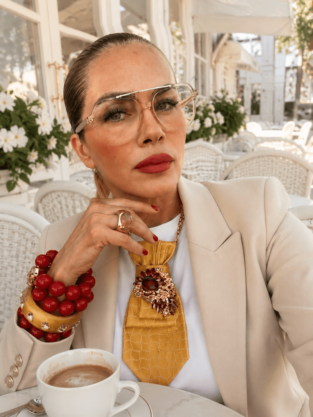 Frau mit heller Brille und rotem Lippenstift sitzt in einem weißen Café. Sie trägt einen beigen Blazer, ein weißes Oberteil, eine gelbe Krawatte mit Schmuckbrosche sowie rote und gelbe Armreifen. Vor ihr steht eine Kaffeetasse auf einem weißen Tisch.