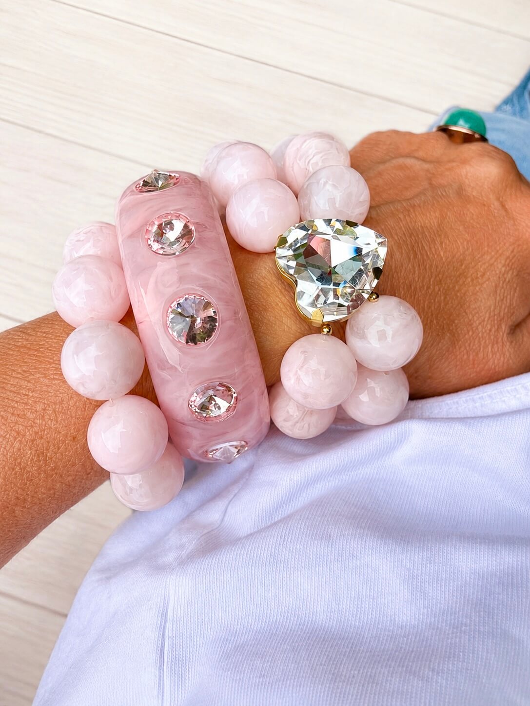 Mehrlagiges Schmuckset in zartem Rosé mit Crystal-Heart-Armband, passendem Bangle mit Schmucksteinen und weiteren Armbändern, getragen am Handgelenk zu weißem Oberteil und Jeans.
