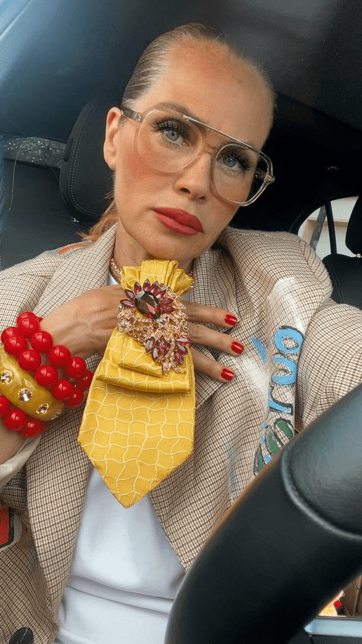 Person mit transparenter Brille trägt eine gelbe Statement-Krawatte mit großer Schmuckbrosche in Rot- und Rosétönen. Kombiniert mit hellem Blazer und roten Armbändern.