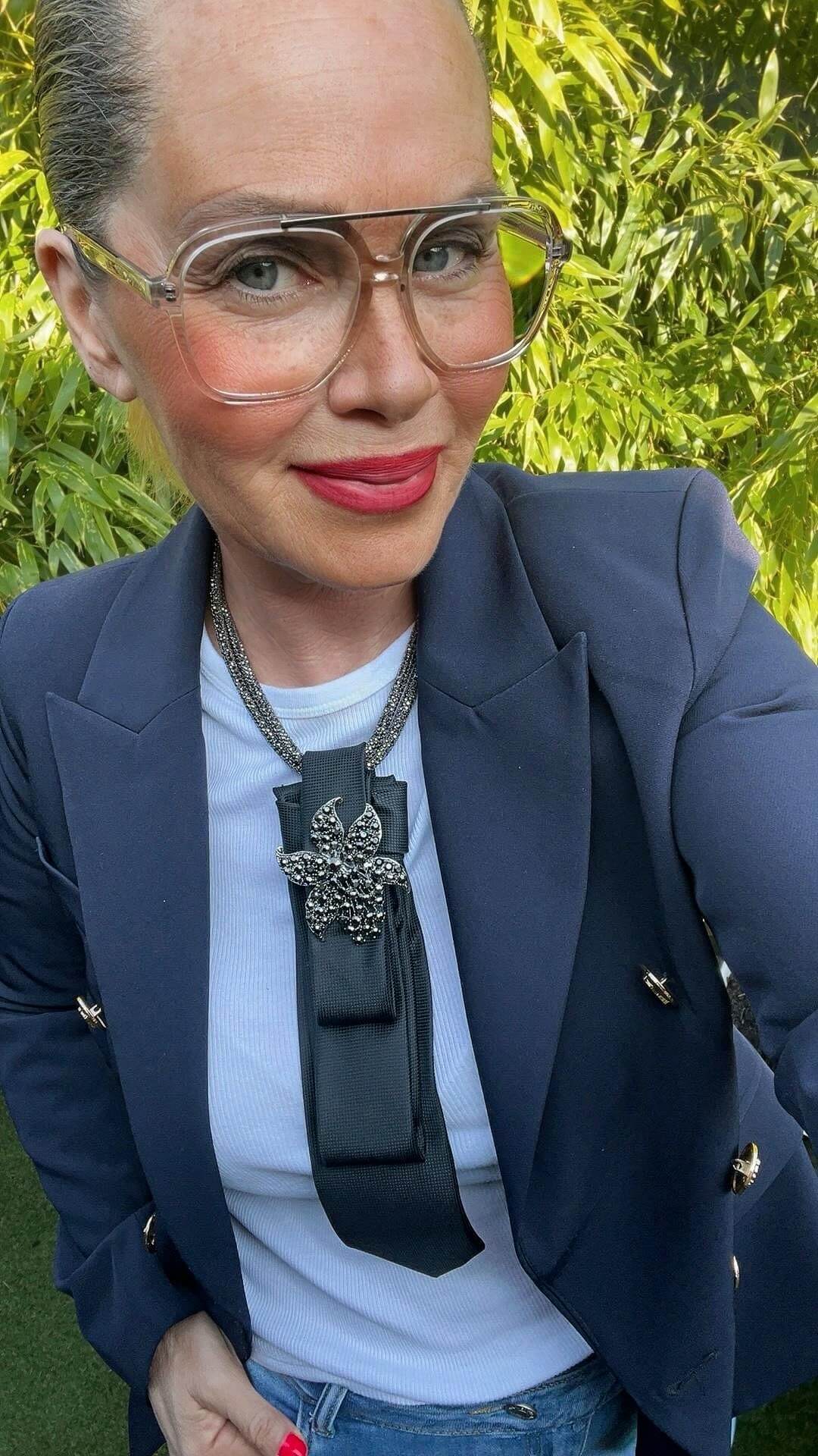 Frau trägt schwarzes Krawatten-Accessoire mit funkelnder Blumenbrosche an weißem Shirt und dunkelblauem Blazer, Nahaufnahme im Freien.
