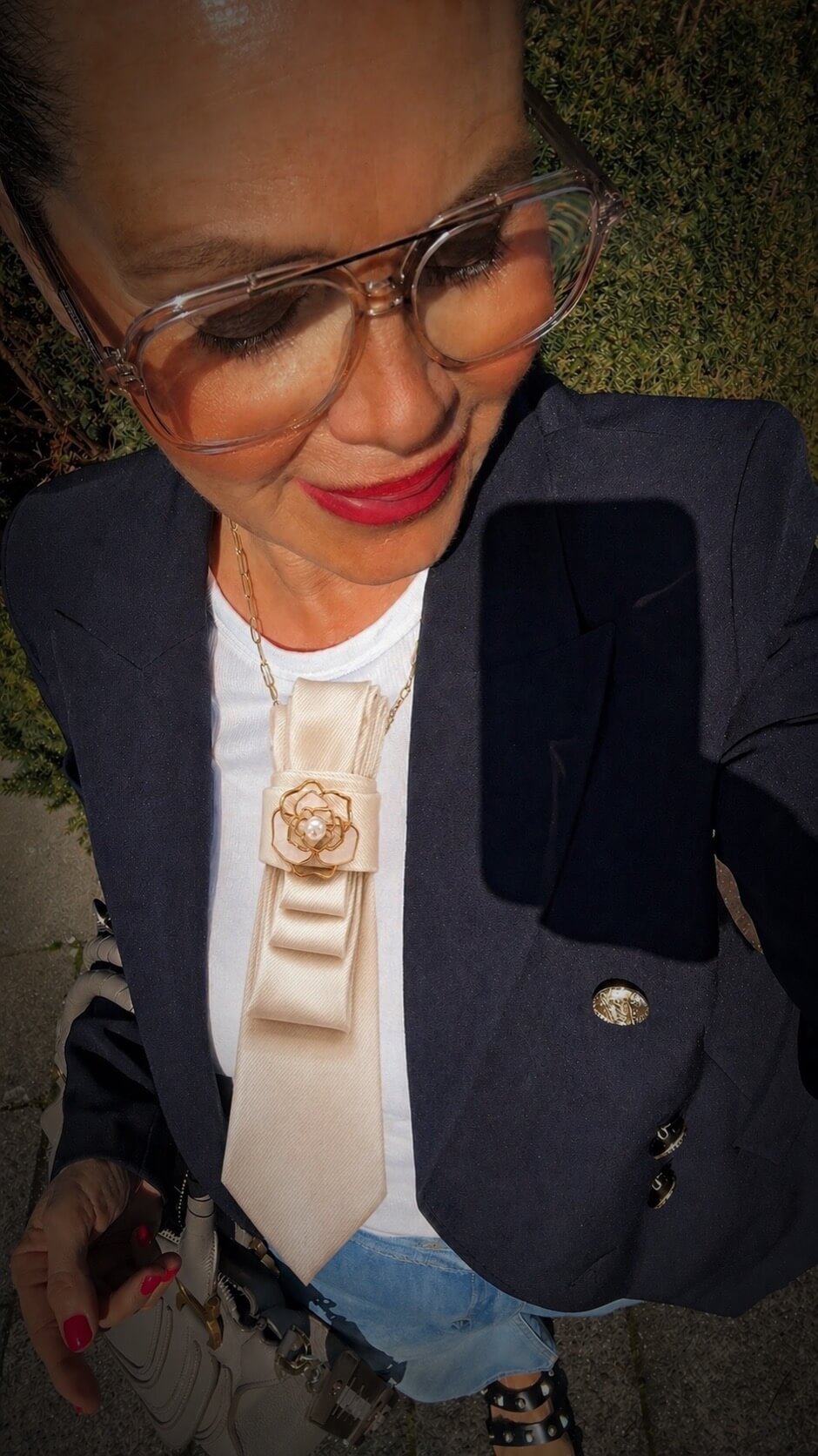 Frau macht ein Selfie im Sonnenlicht, trägt eine große transparente Brille, dunkelblauen Blazer über weißem Oberteil sowie eine cremefarbene Krawatte mit floraler Brosche, dazu rote Nägel und eine Handtasche.