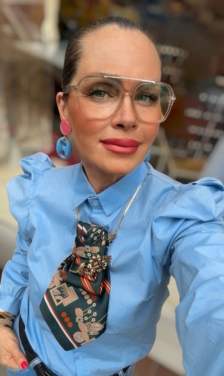 Person trägt eine hellblaue Bluse und eine dunkelgrüne Krawatte mit grafischem Muster, kombiniert mit dekorativem Schmuckelement, als Nahaufnahme im Brustbereich dargestellt.