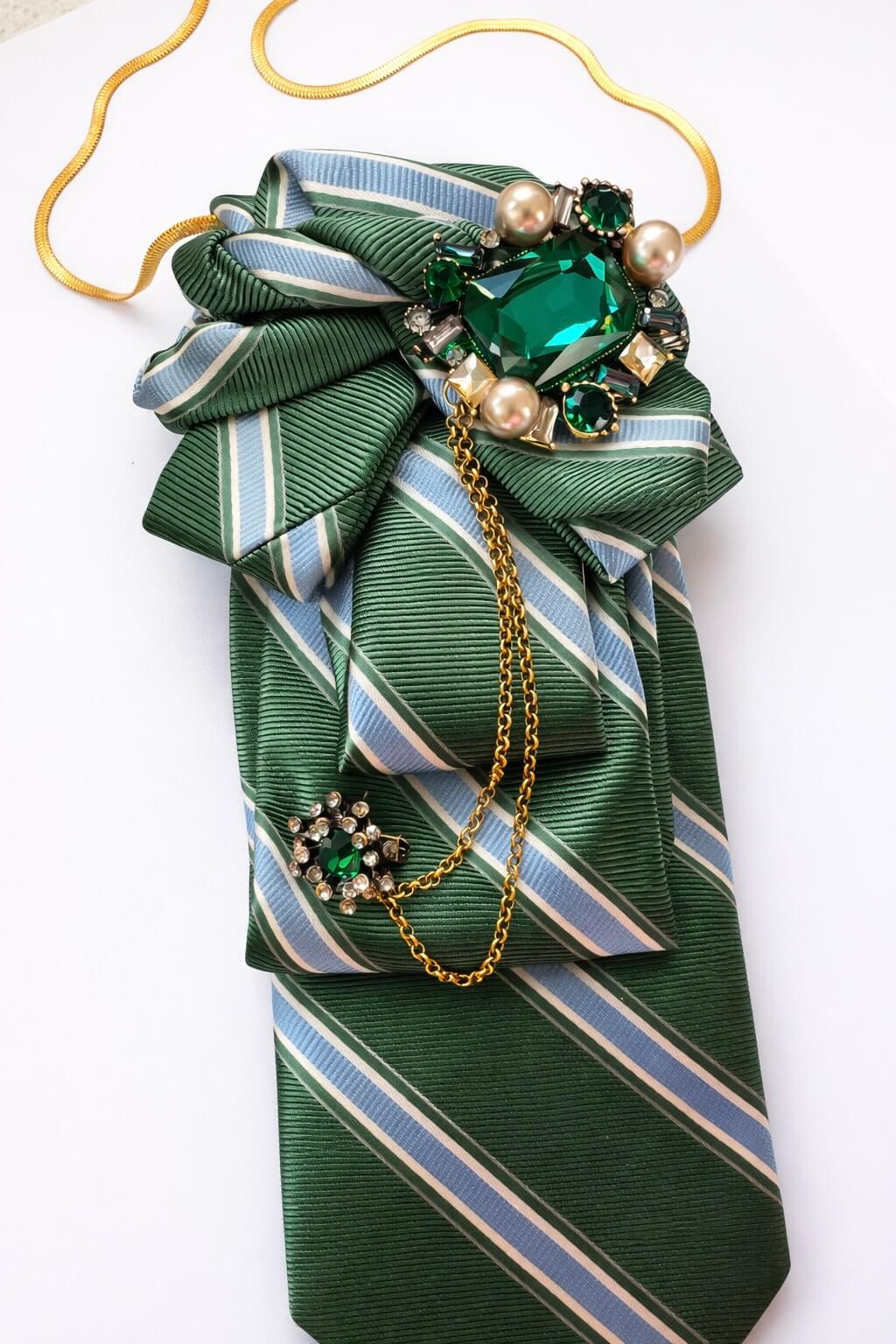 Grüne gestreifte Krawatte mit dekorativer Schleife, verziert mit einer großen grünen Schmuckbrosche, Perlen und einer goldfarbenen Kette, auf hellem Hintergrund.