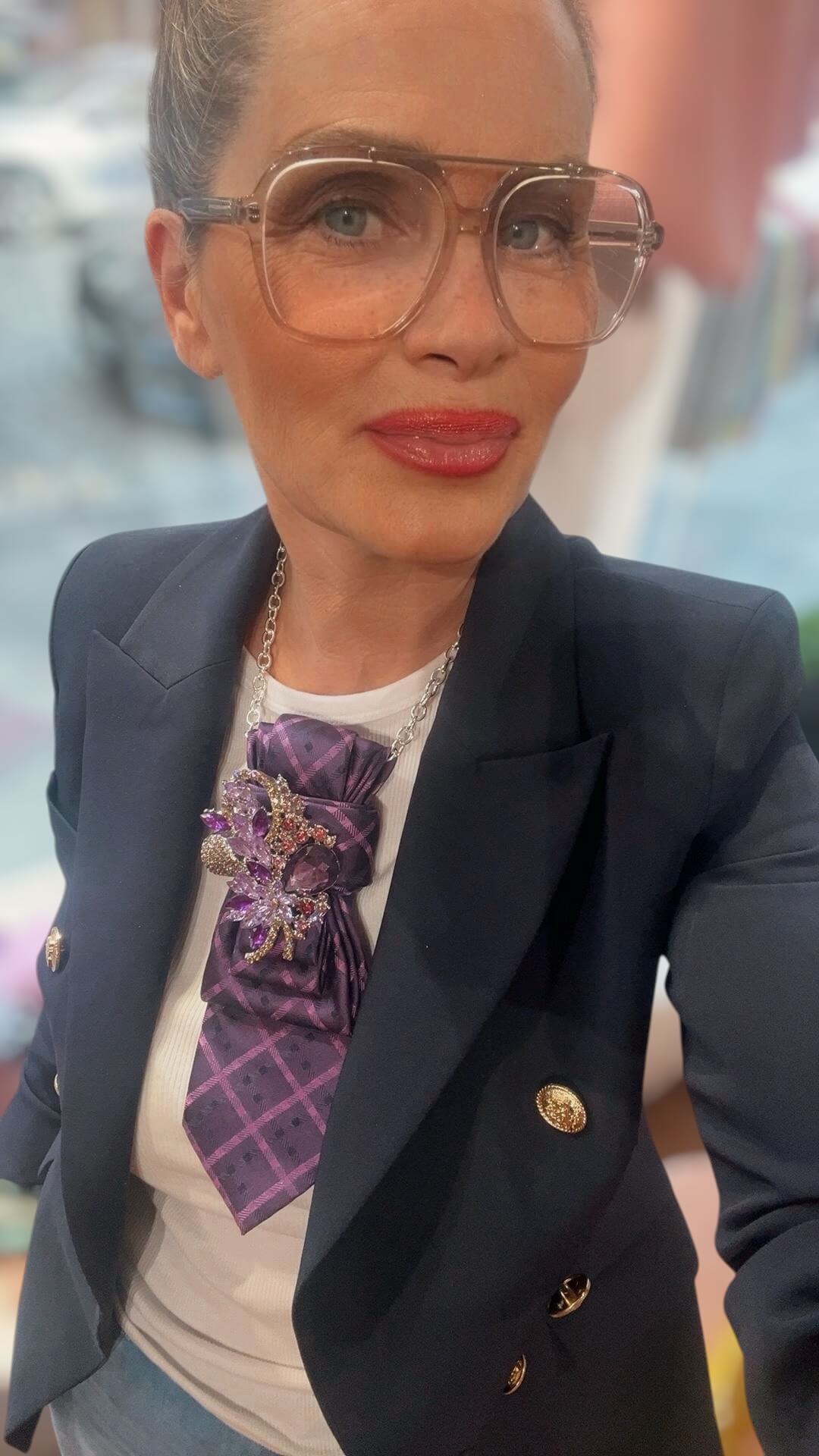 Nahaufnahme einer Frau mit Brille, dunkelblauem Blazer und weißem Oberteil, trägt eine lilafarbene Krawatte mit verzierter Brosche als Blickfang.