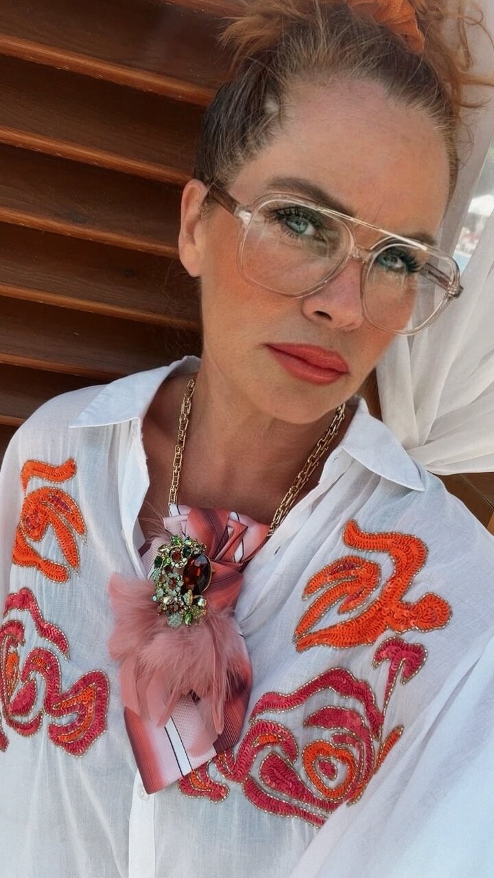 Frau mit heller Brille und hochgesteckten Haaren trägt eine weiß-transparente Bluse mit orange- und pinkfarbenen Stickereien. Am Hals befindet sich eine rosafarbene Krawatte mit Brosche und rosa Federdetail an einer goldfarbenen Kette. Die Aufnahme zeigt die Person von der Brust aufwärts vor einer Holzverkleidung.