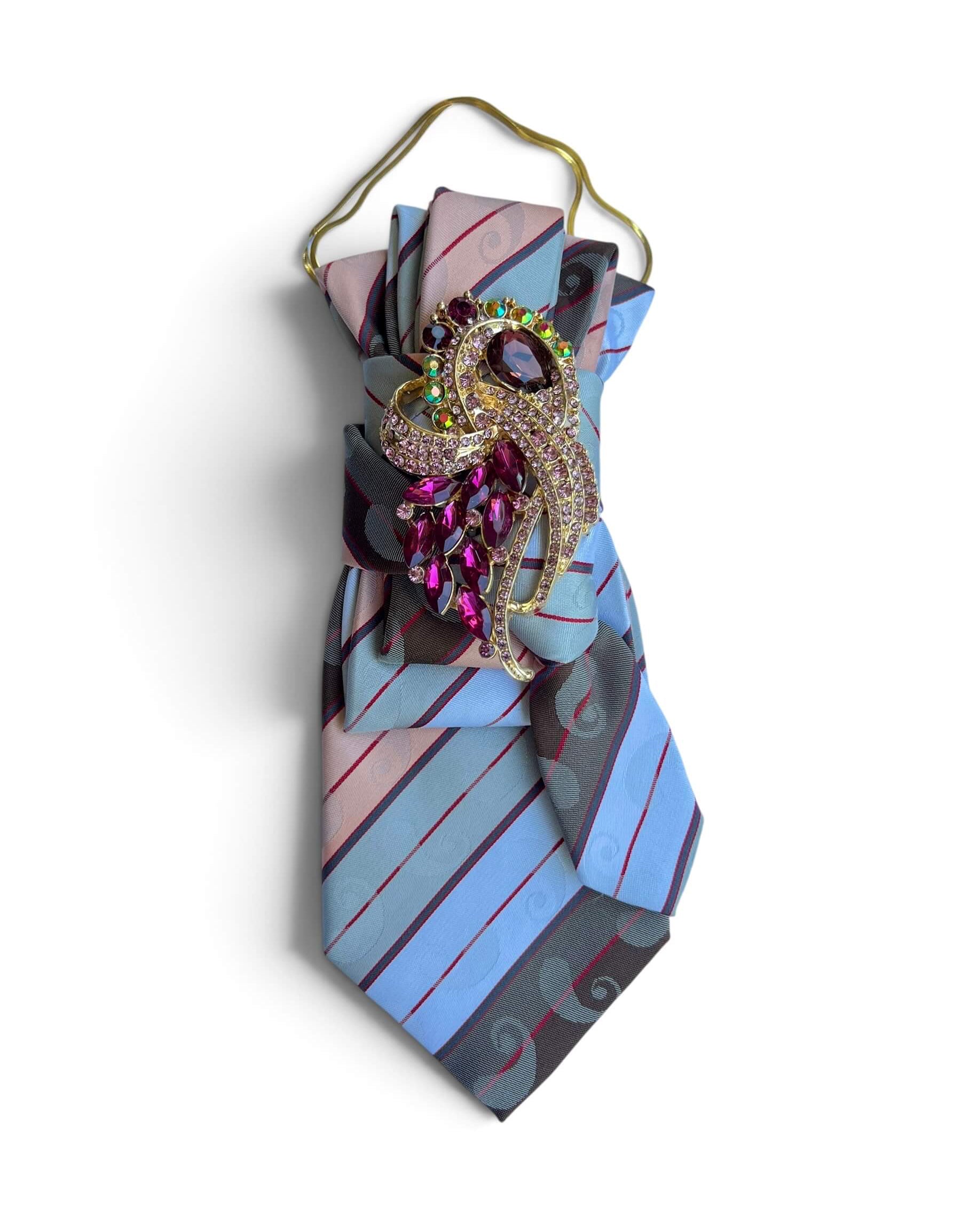 Hellblaue Statement-Krawatte mit Ornamentmuster, pinkfarbener Schmuckbrosche und goldfarbener Kette.