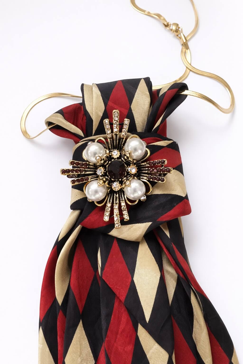 Krawatte mit Rautenmuster in Rot, Schwarz und Beige, kombiniert mit goldfarbener Kette und dekorativem Schmuckelement mit Perlen und Strass, frontal auf hellem Hintergrund dargestellt. Im Detail.