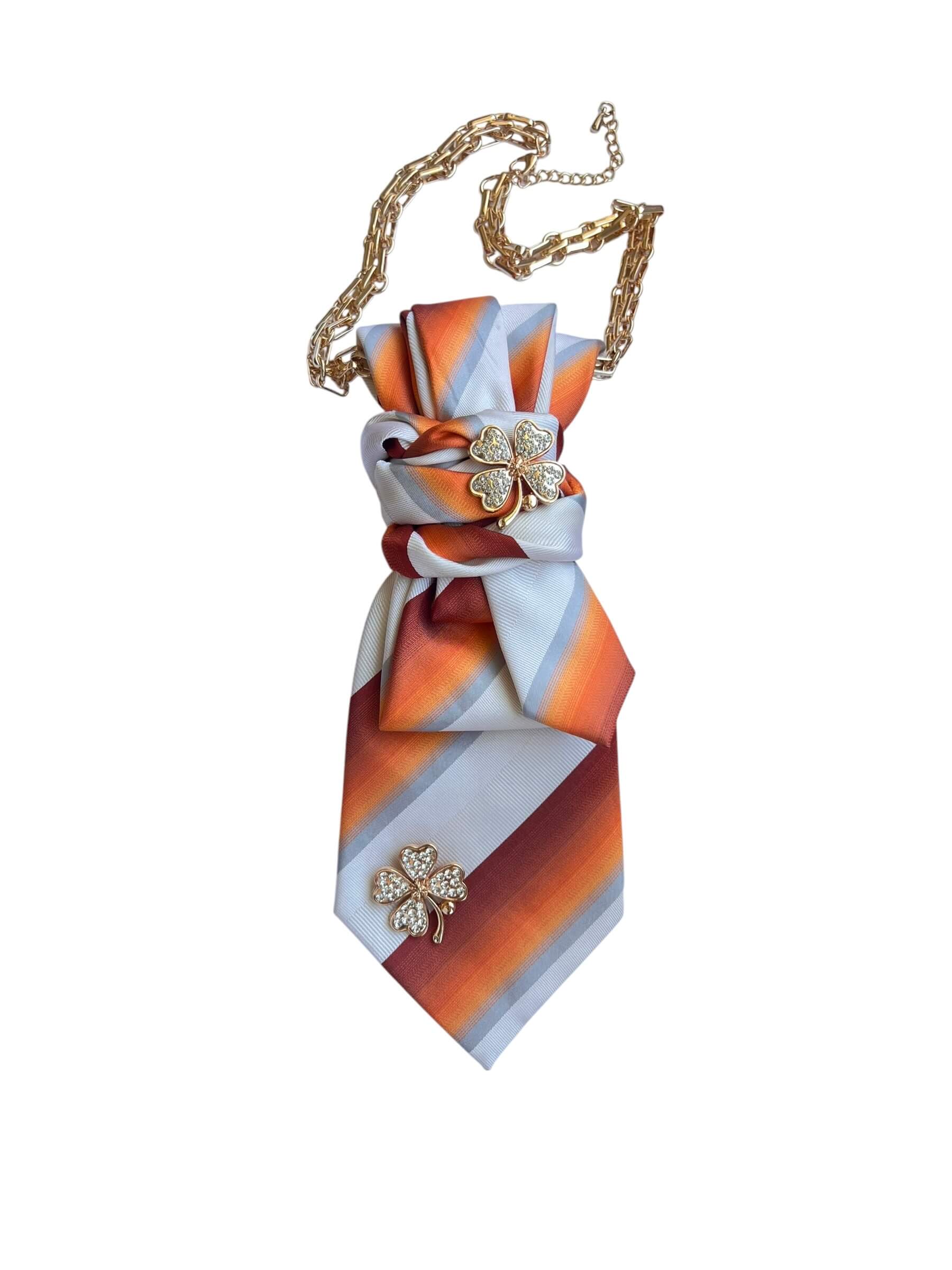 Seidenkrawatte mit diagonalen Streifen in Orange, Weiß und Rot, kombiniert mit einer Kleeblatt-Brosche und goldfarbener Kette, freigestellt auf hellem Hintergrund.