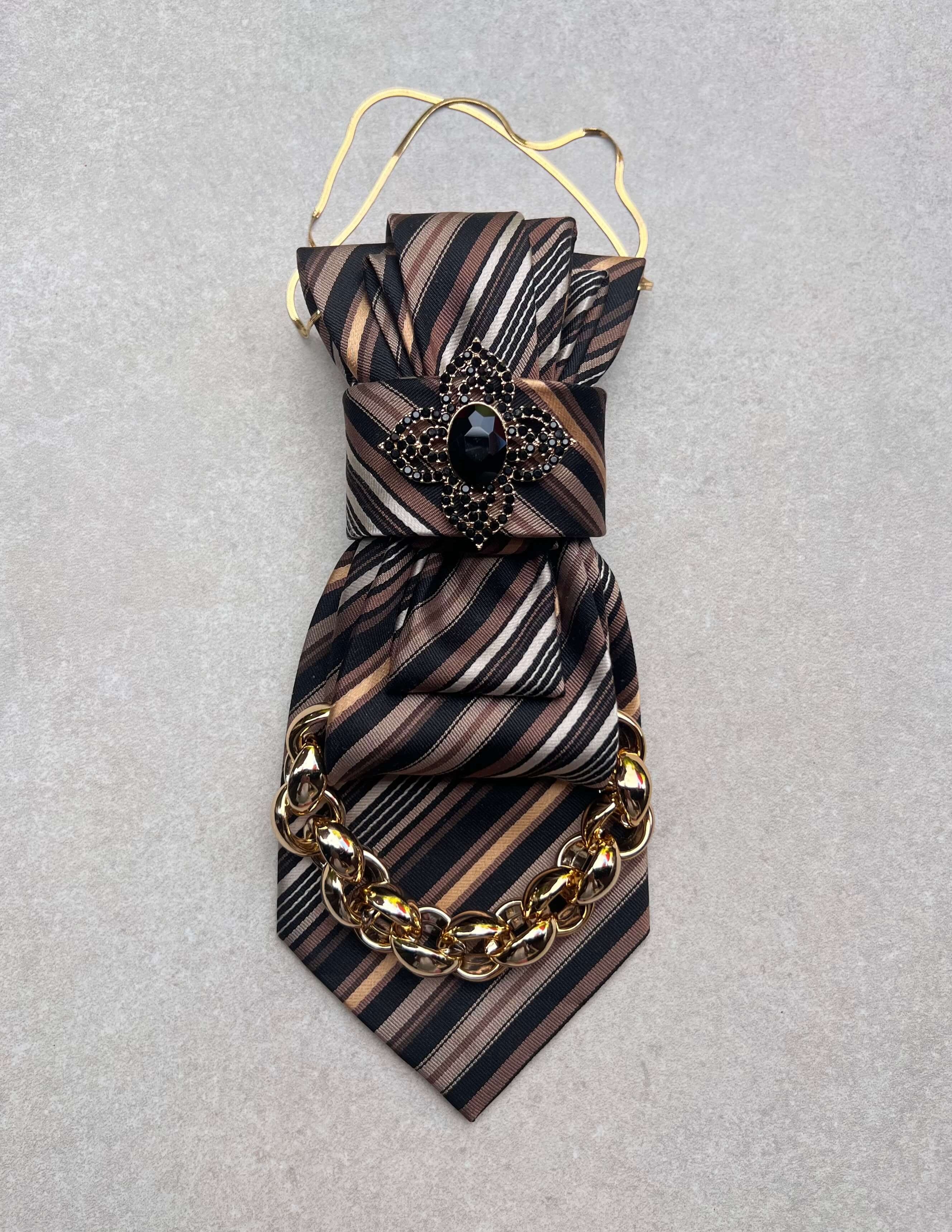 Gestaltete Krawattenkette aus gestreiftem Stoff in Schwarz-, Taupe- und Brauntönen mit goldfarbener Kette und schwarzer Schmuckbrosche auf hellem Hintergrund.