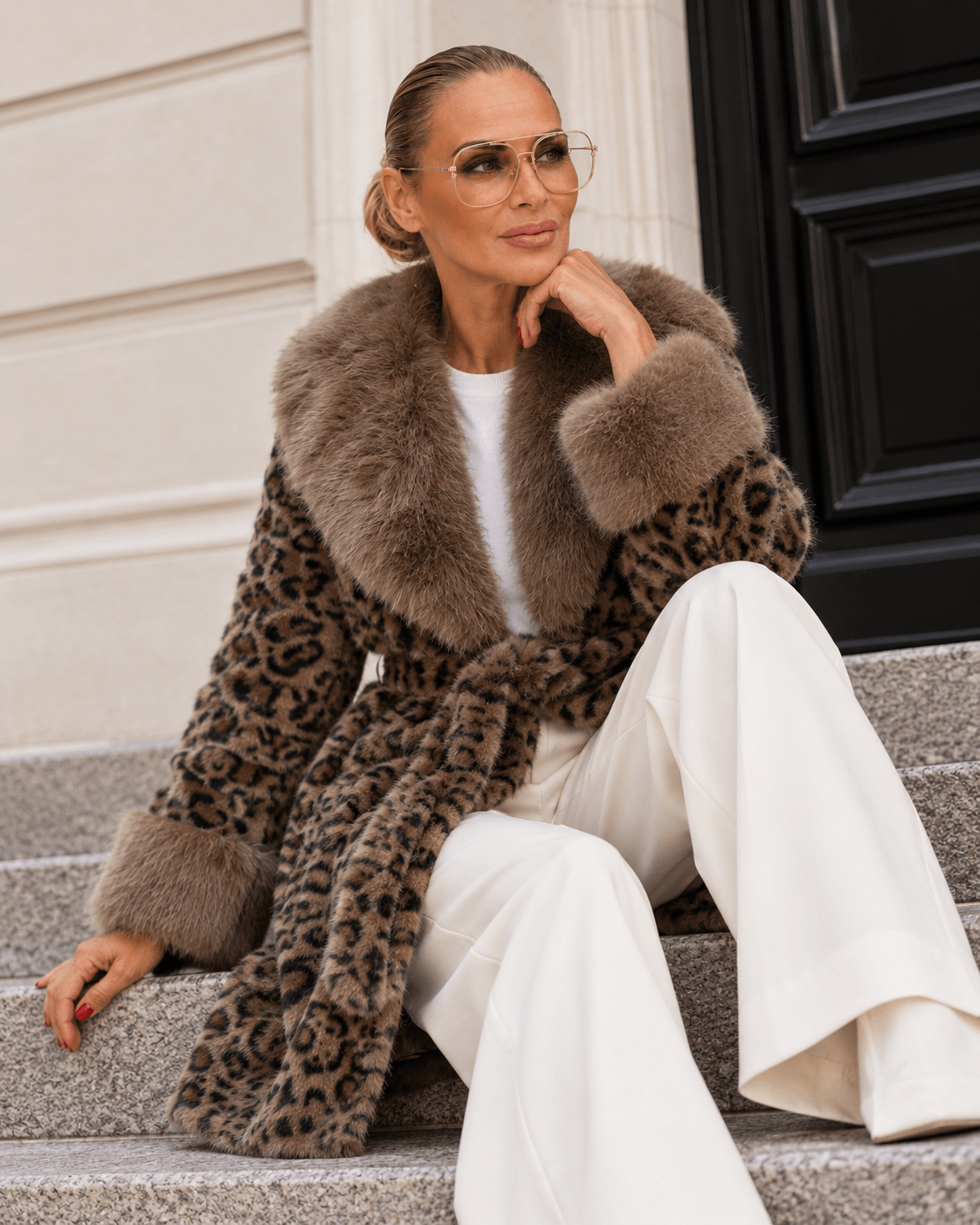     Model trägt die Leo Fake Fur Jacket von Casapari mit Gürtel und weißer Marlene, sitzend auf einer Treppe.