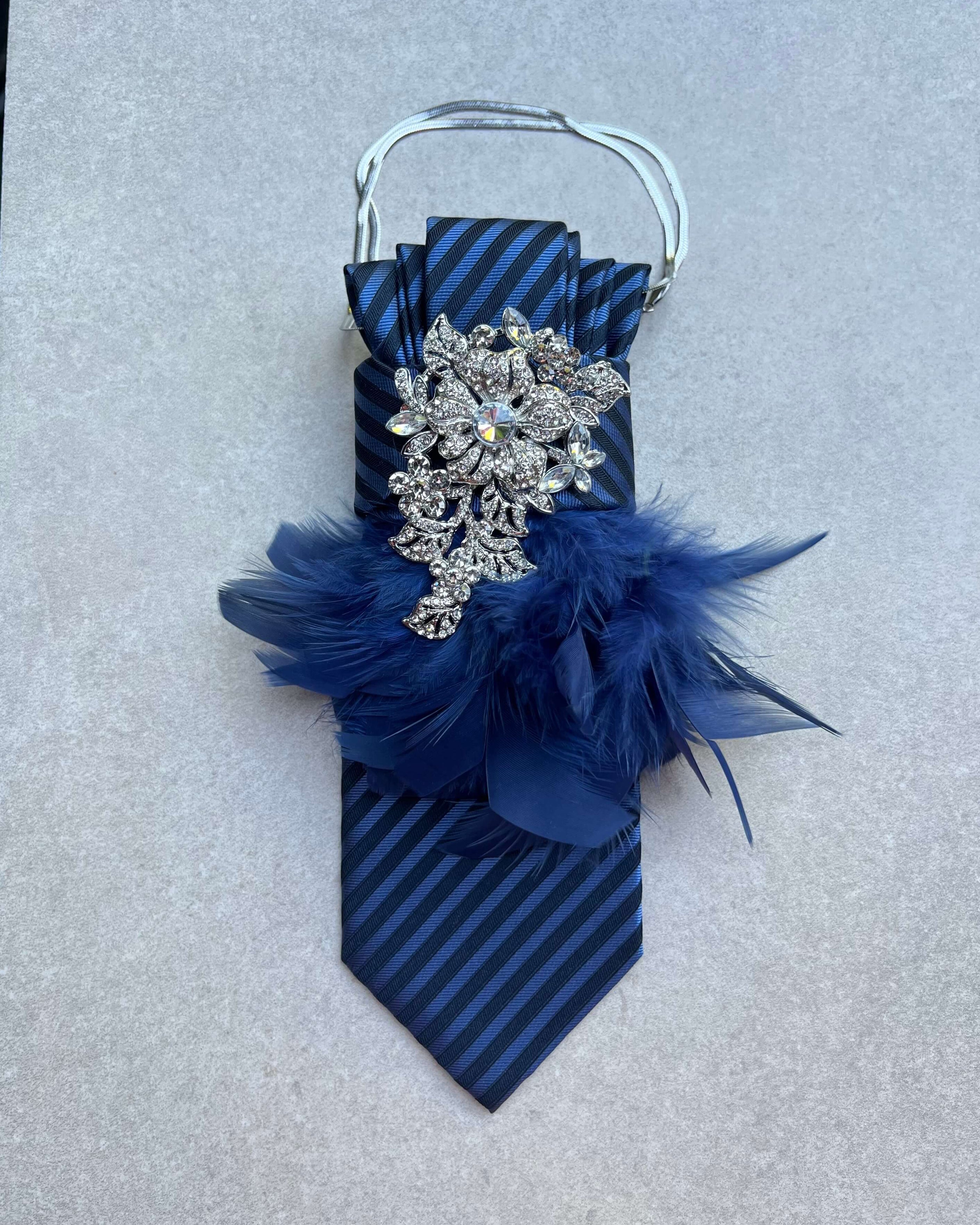 Dunkelblaue Statement-Krawatte mit Streifenmuster, silberfarbener Schmuckbrosche, blauen Federn und silberfarbener Kette auf heller Steinoberfläche.