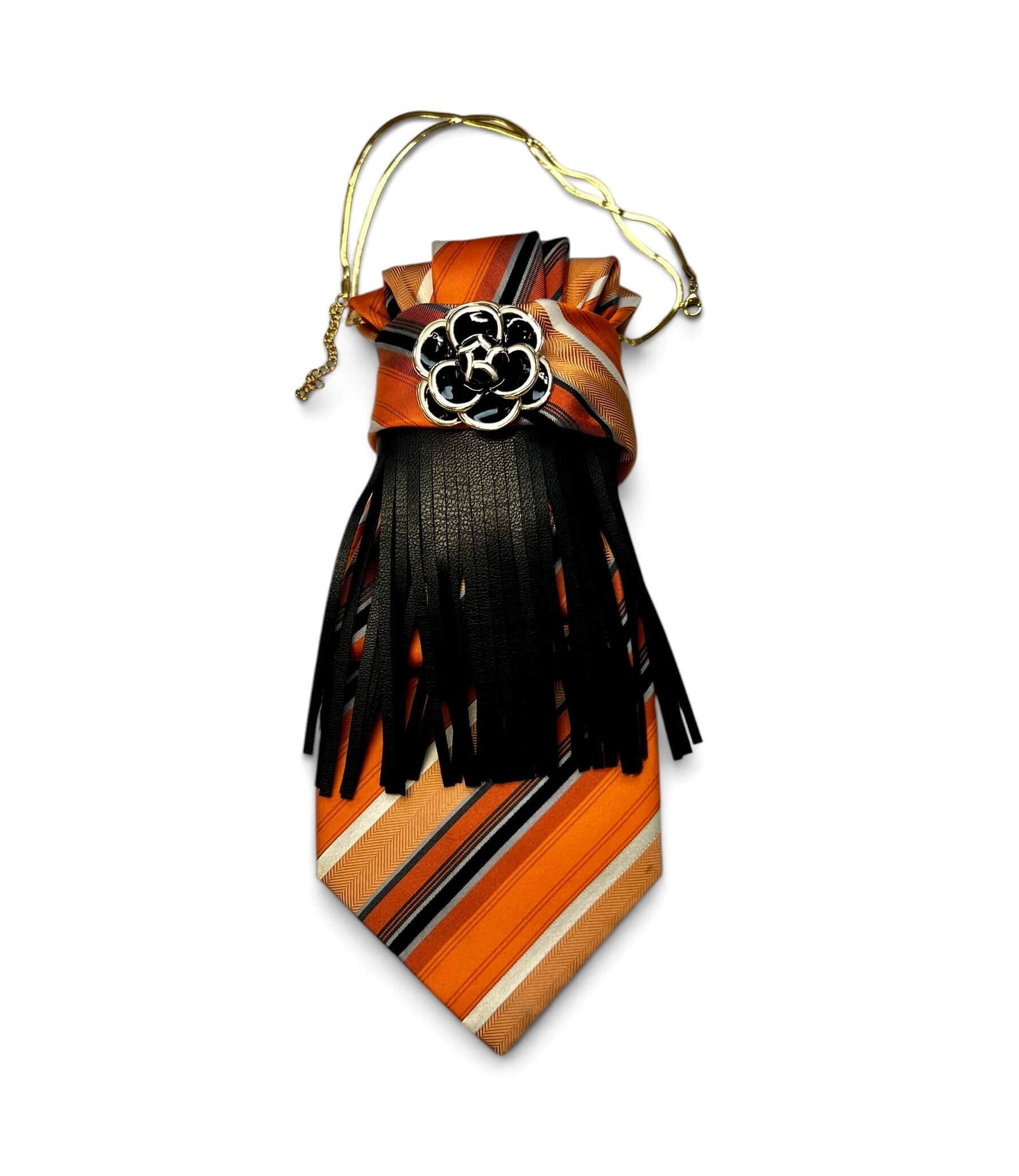 Gestreifte Krawatte in Orange, Beige und Schwarz mit schwarzen Lederfransen, Blütenbrosche und goldfarbener Kette auf hellem Hintergrund.