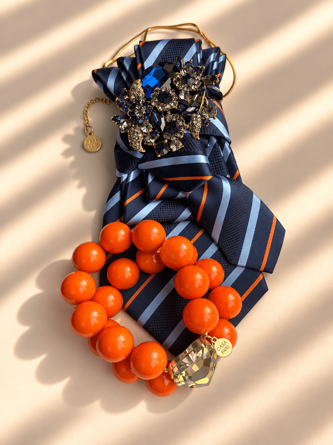 Orangenes Armband mit goldenem Herz, blaue Krawatte mit blau orangenen Streifen, große Brosche,kette auf hellem Hintergrund fotografiert