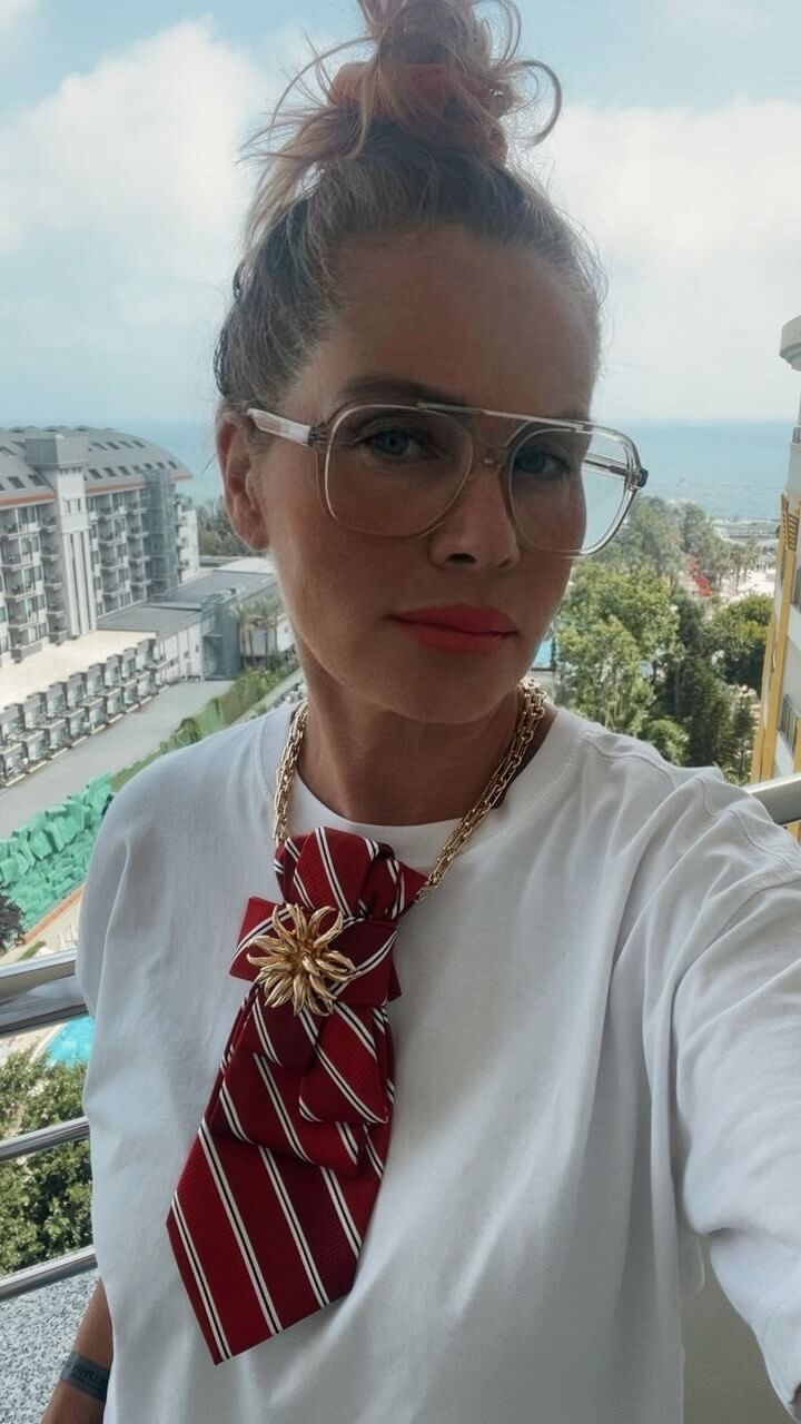 Frau mit heller Brille und hochgesteckten Haaren trägt ein weißes Oberteil. Am Hals befindet sich eine rote gestreifte Krawatte mit goldfarbener Brosche an einer goldfarbenen Kette. Die Person steht auf einem Balkon, im Hintergrund sind Gebäude und Himmel zu sehen.