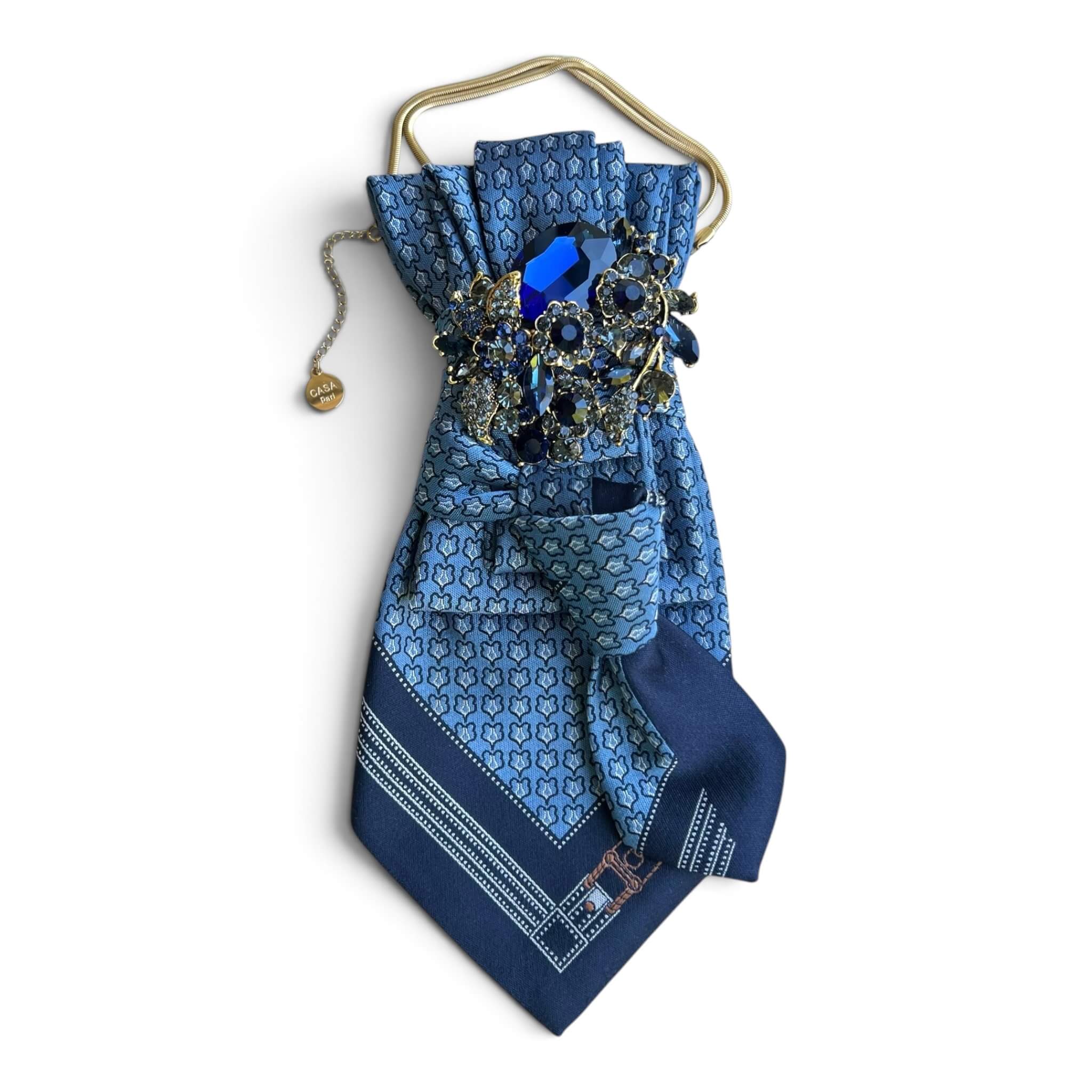 Blau gemusterte CASAPARI Seidenkrawatte mit dunkelblauen Partien, goldfarbener Kette und großer Schmuckbrosche mit blauen Kristallsteinen.