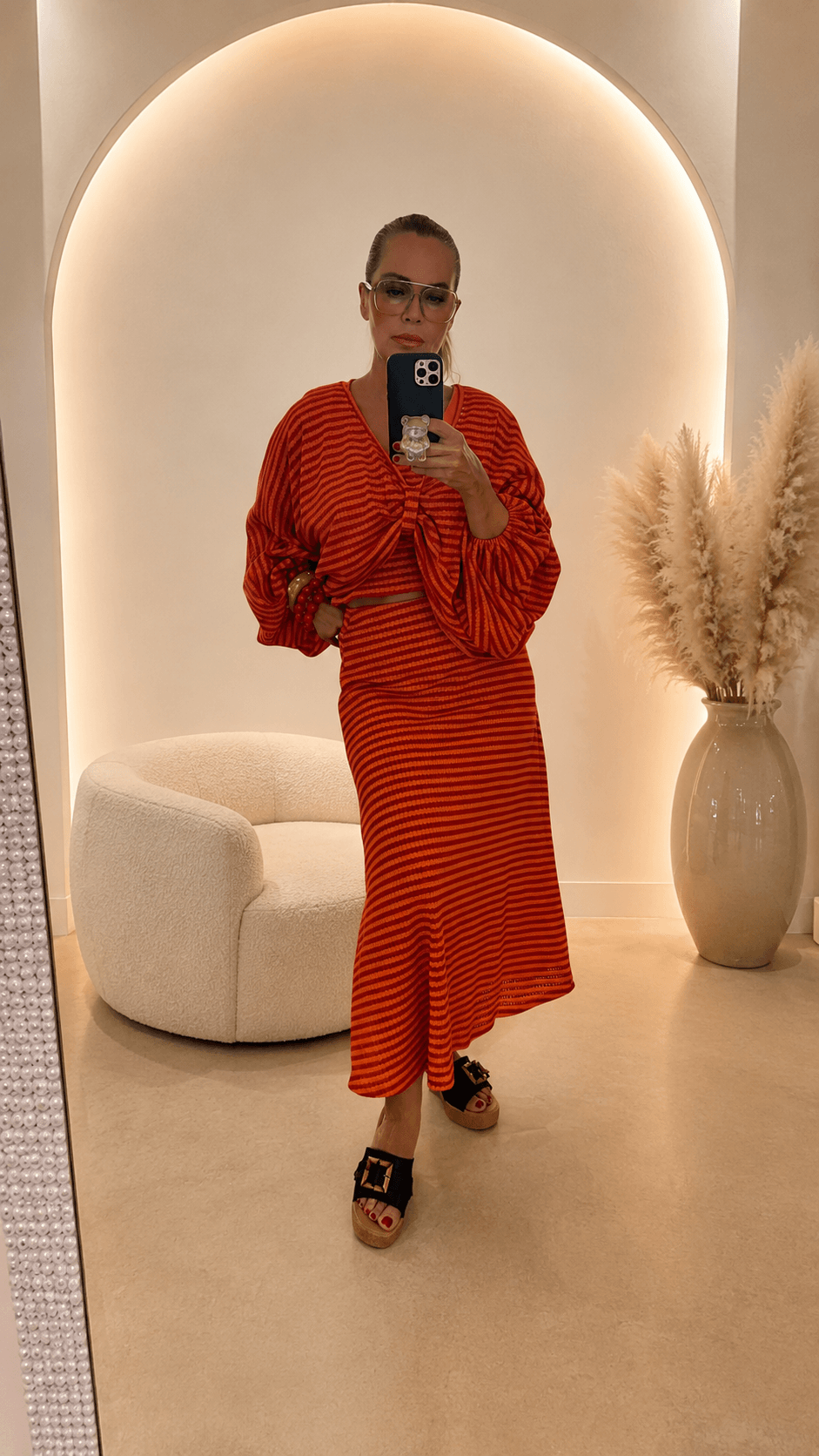 Frau trägt ein orange-rotes, fein gestreiftes Set mit Tank-Crop-Top, Fledermaus-Pullover mit Raffung vorne und langem Rock in A-Linie. Dazu schwarze Plateau-Sandalen und farblich passende Armreifen.