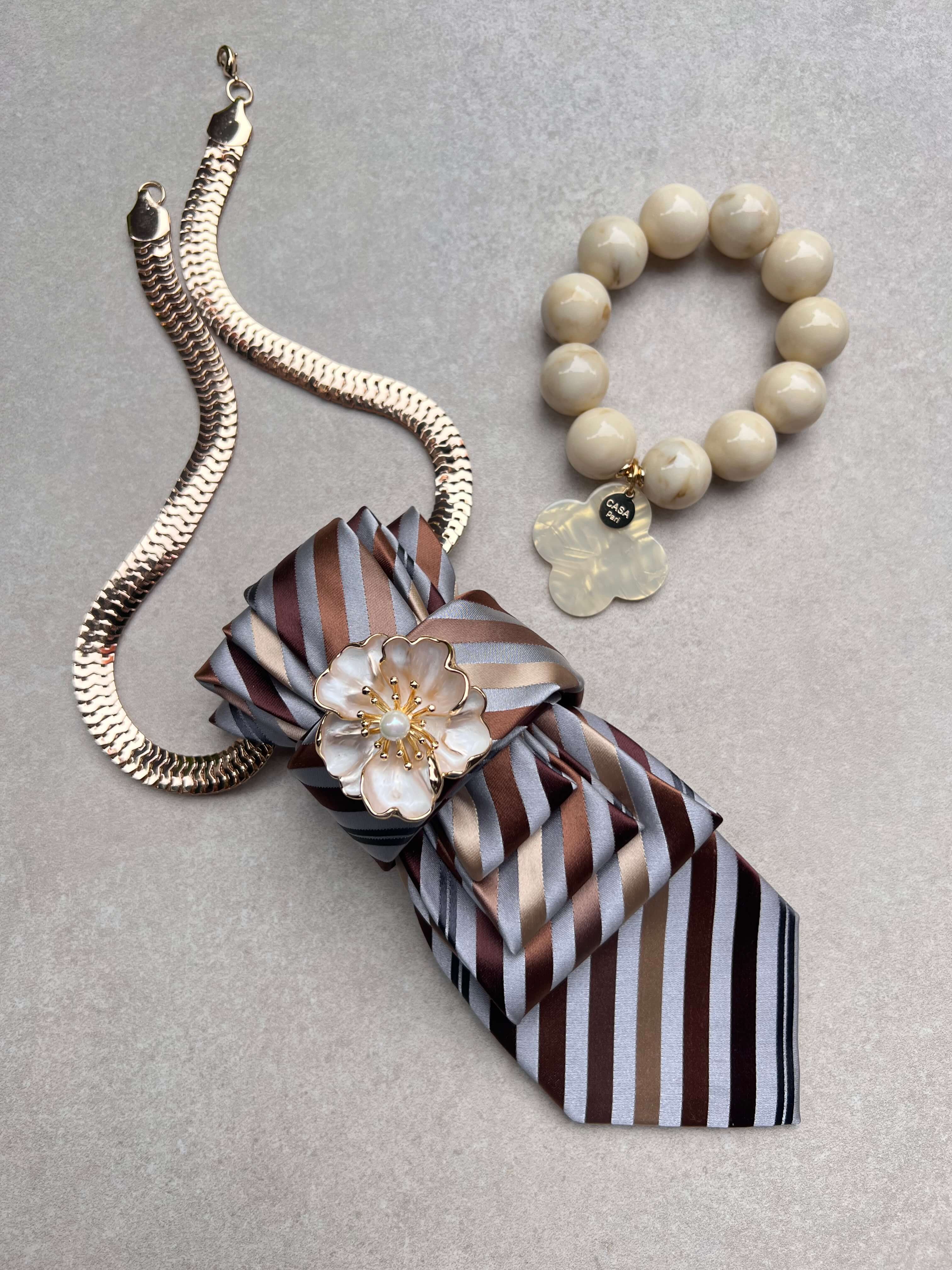 Kombiniertes Set aus gestreifter Krawattenkette mit goldfarbener Kette und Blütenbrosche sowie passendem Armband mit Kleeblatt-Anhänger in Sand-, Creme- und Brauntönen.