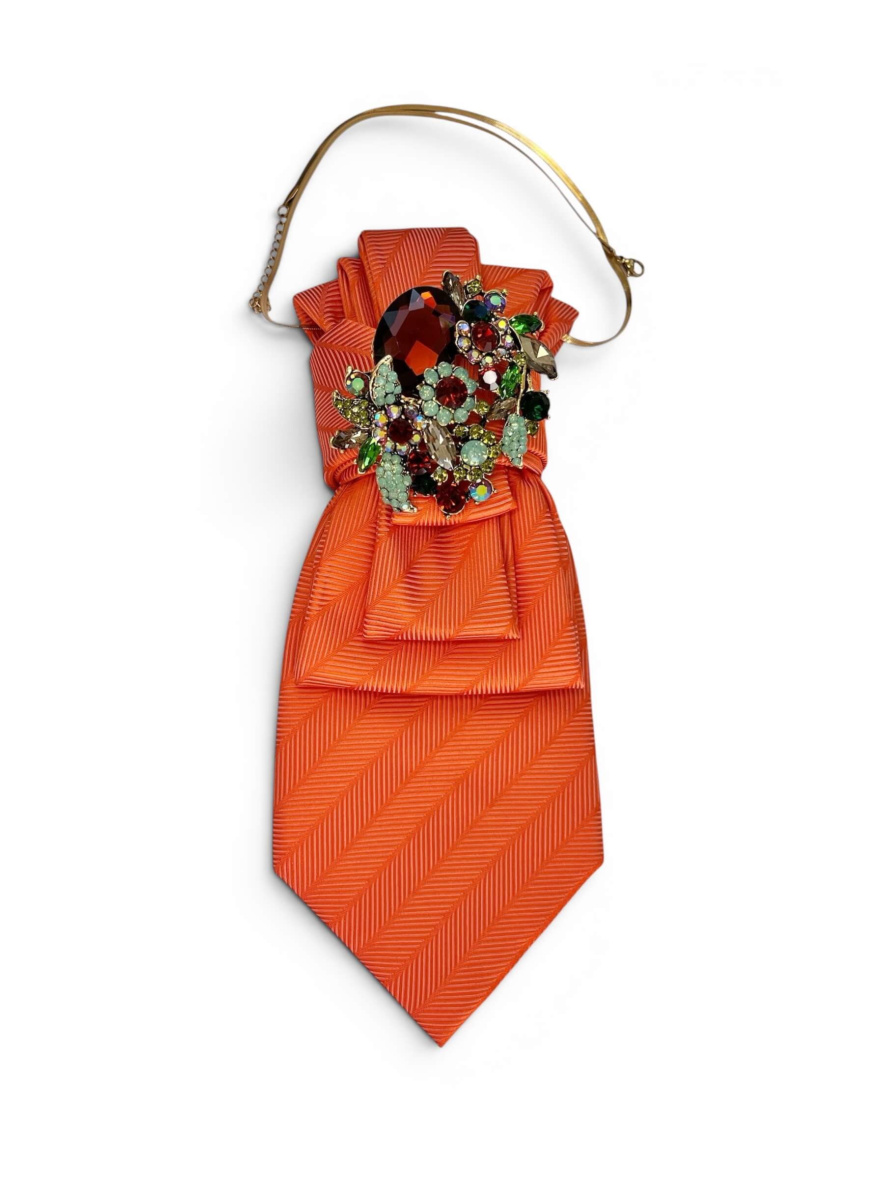 Orange Vintage-Krawatte mit diagonaler Struktur, dekorativ gebunden mit mehrfarbiger Schmuckbrosche mit roten, grünen und hellblauen Steinen vor neutralem Hintergrund