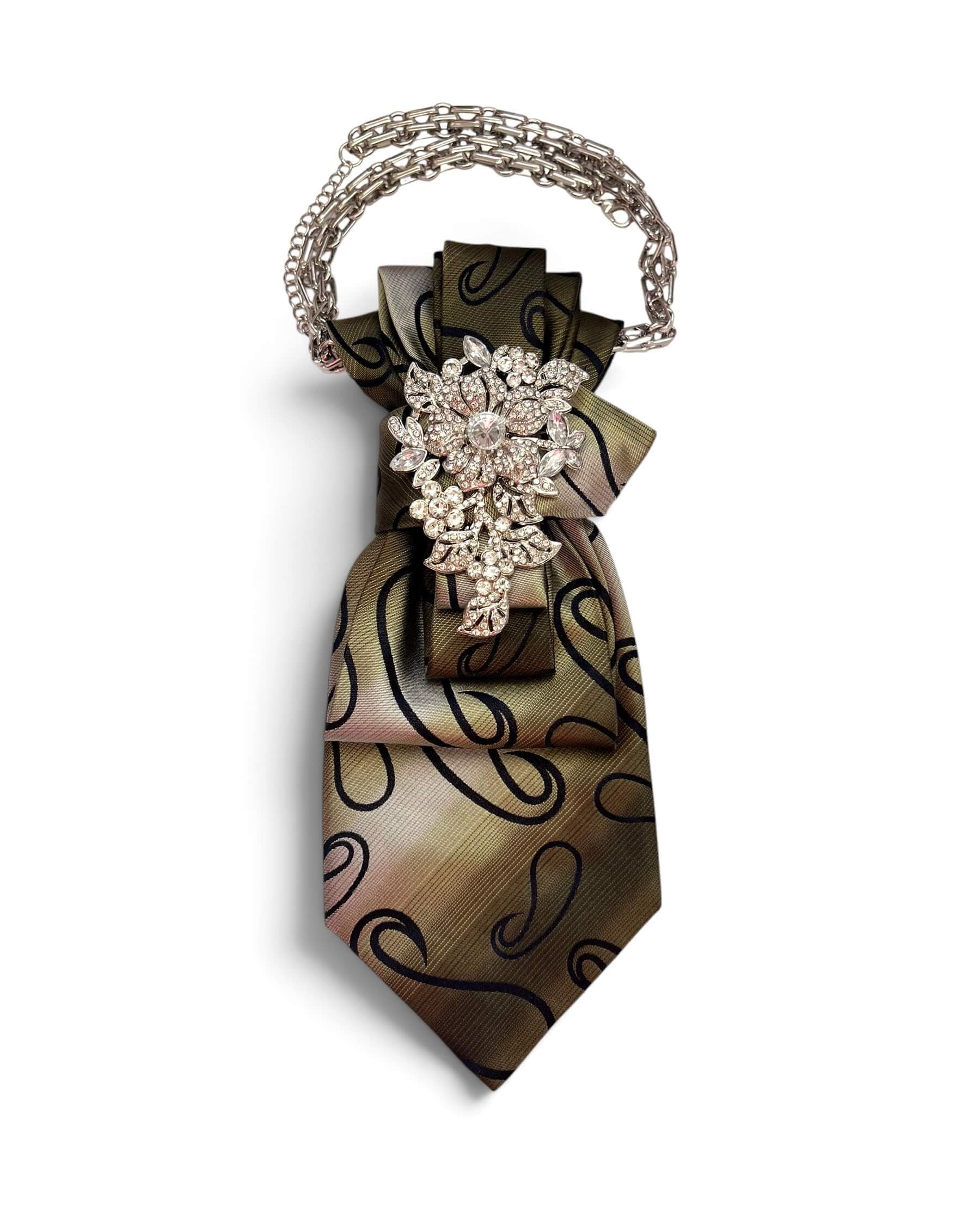 Seidenkrawatte in Taupe- und Mokkatönen mit dunklem Muster als Schmuckkette mit silberfarbener Kette und kristallbesetzter Brosche vor neutralem Hintergrund.