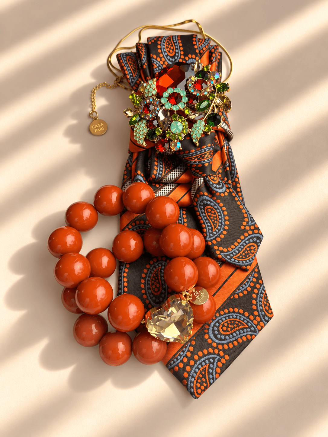 Silk Affair Armband Spiced Orange von Casapari mit großen orangefarbenen Perlen, facettiertem Crystal Heart und goldenem Anhänger, kombiniert mit braun-oranger Statement-Krawatte.