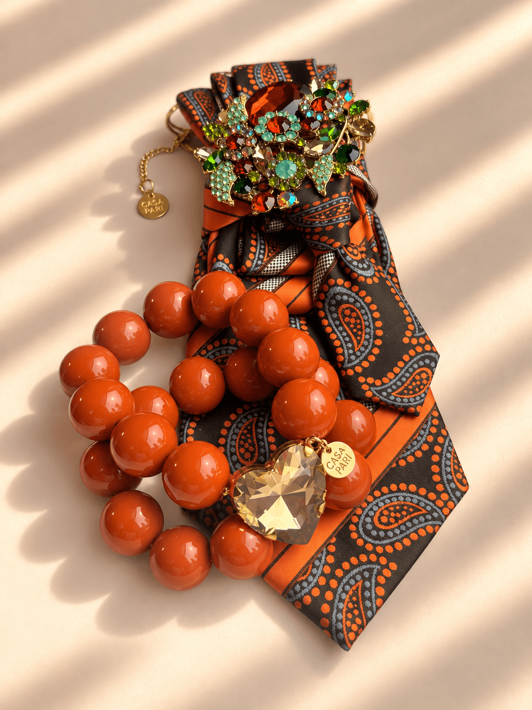 Silk Affair Armband Spiced Orange von Casapari mit großen orangefarbenen Perlen, facettiertem Crystal Heart und goldenem Anhänger, kombiniert mit braun-oranger Statement-Krawatte.