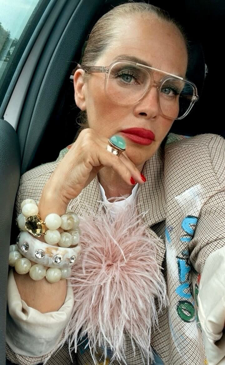 Frau mit transparenter Brille, roten Lippen und gemustertem Blazer sitzt im Auto und präsentiert mehrere helle Armreifen, einen Ring mit türkisfarbenem Stein und eine rosafarbene Federbrosche.
