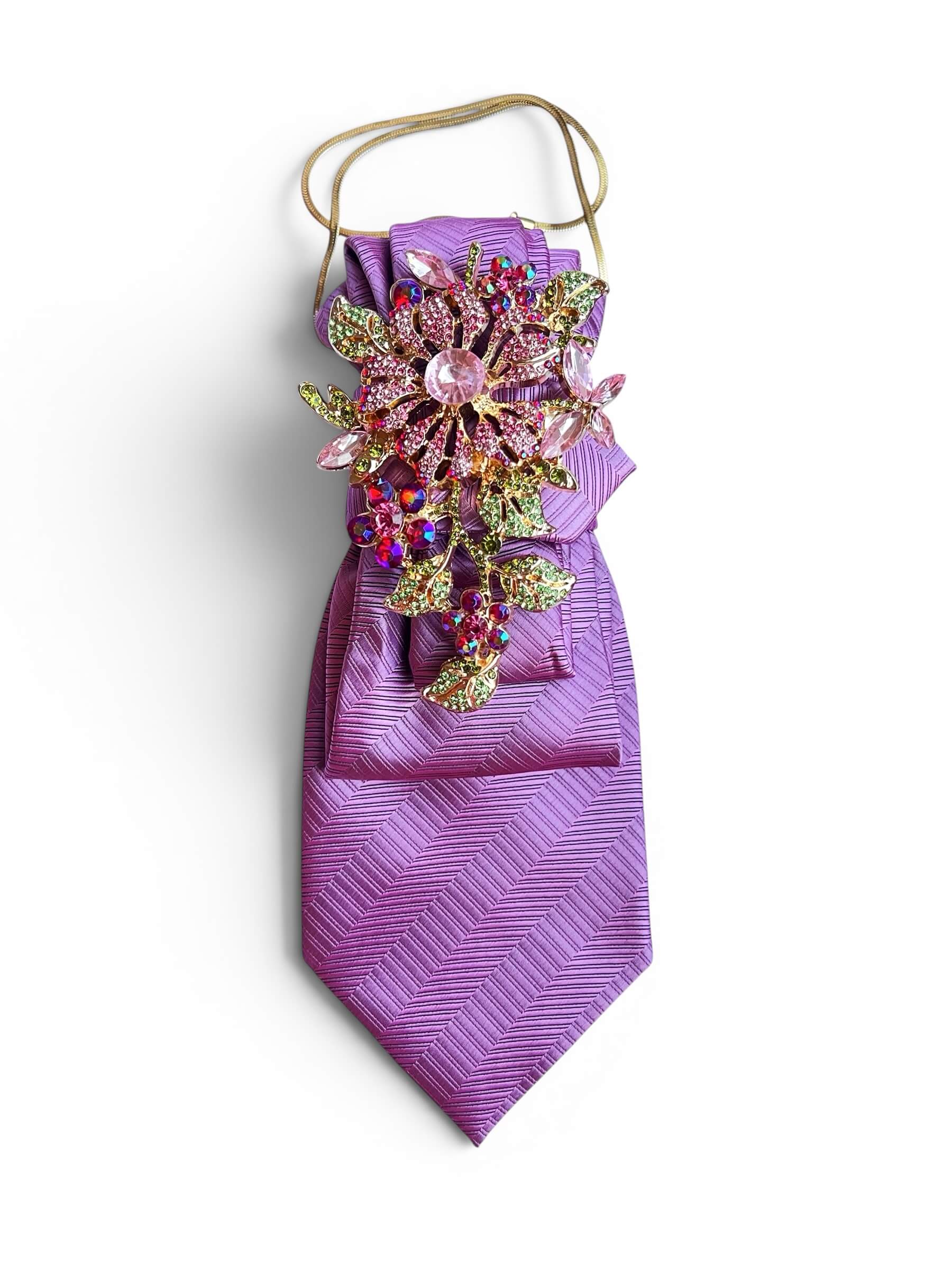 Rosa-lila Vintage-Krawatte mit diagonaler Struktur, dekorativer Schmuckbrosche mit rosa und mehrfarbigen Steinen sowie feiner goldfarbener Kette auf hellem Hintergrund