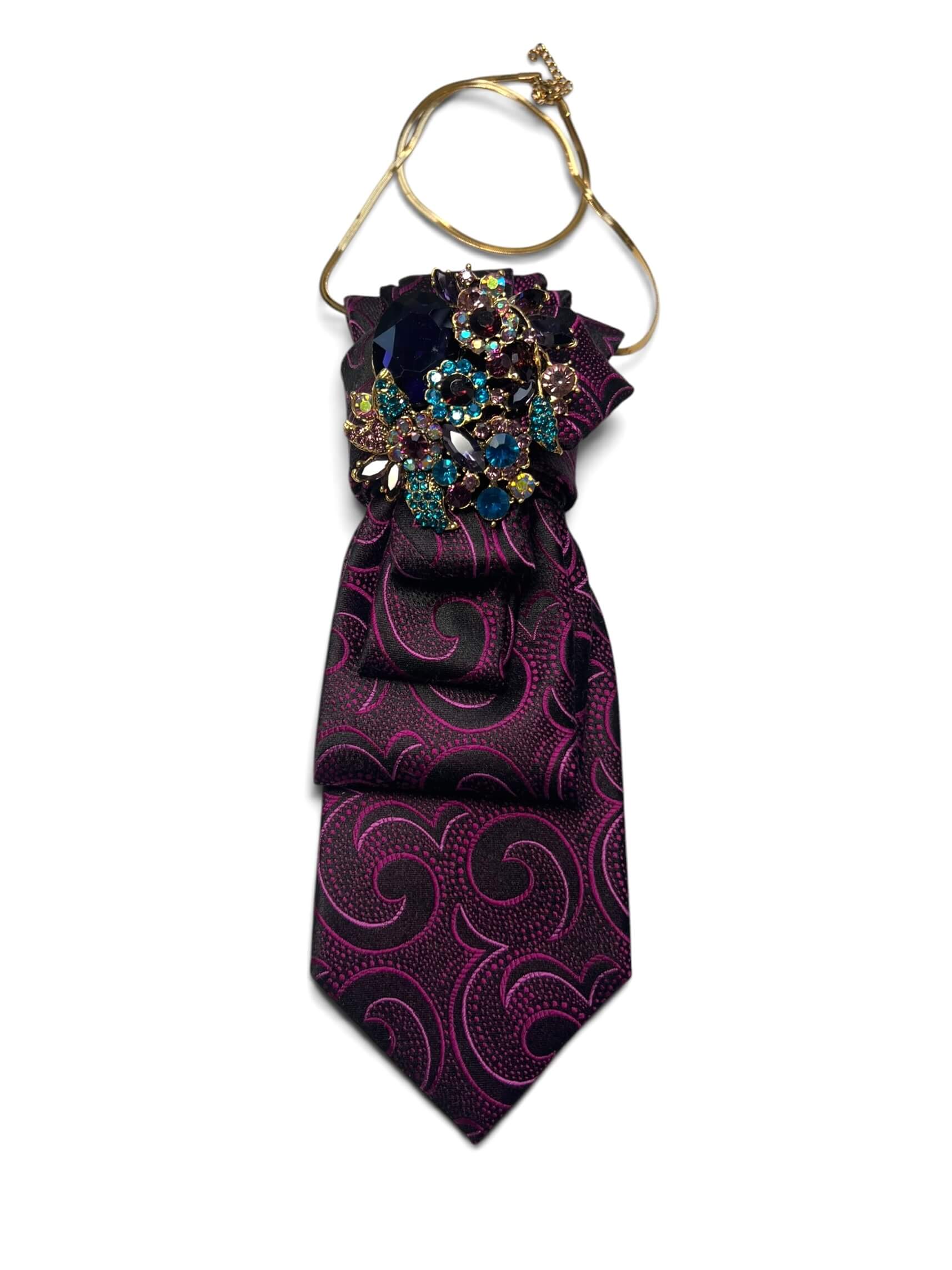 Vintage-Krawatte in Schwarz mit pinkfarbenem Ornamentmuster, dekorativ drapiert mit farbiger Schmuckbrosche und goldfarbener Kette.