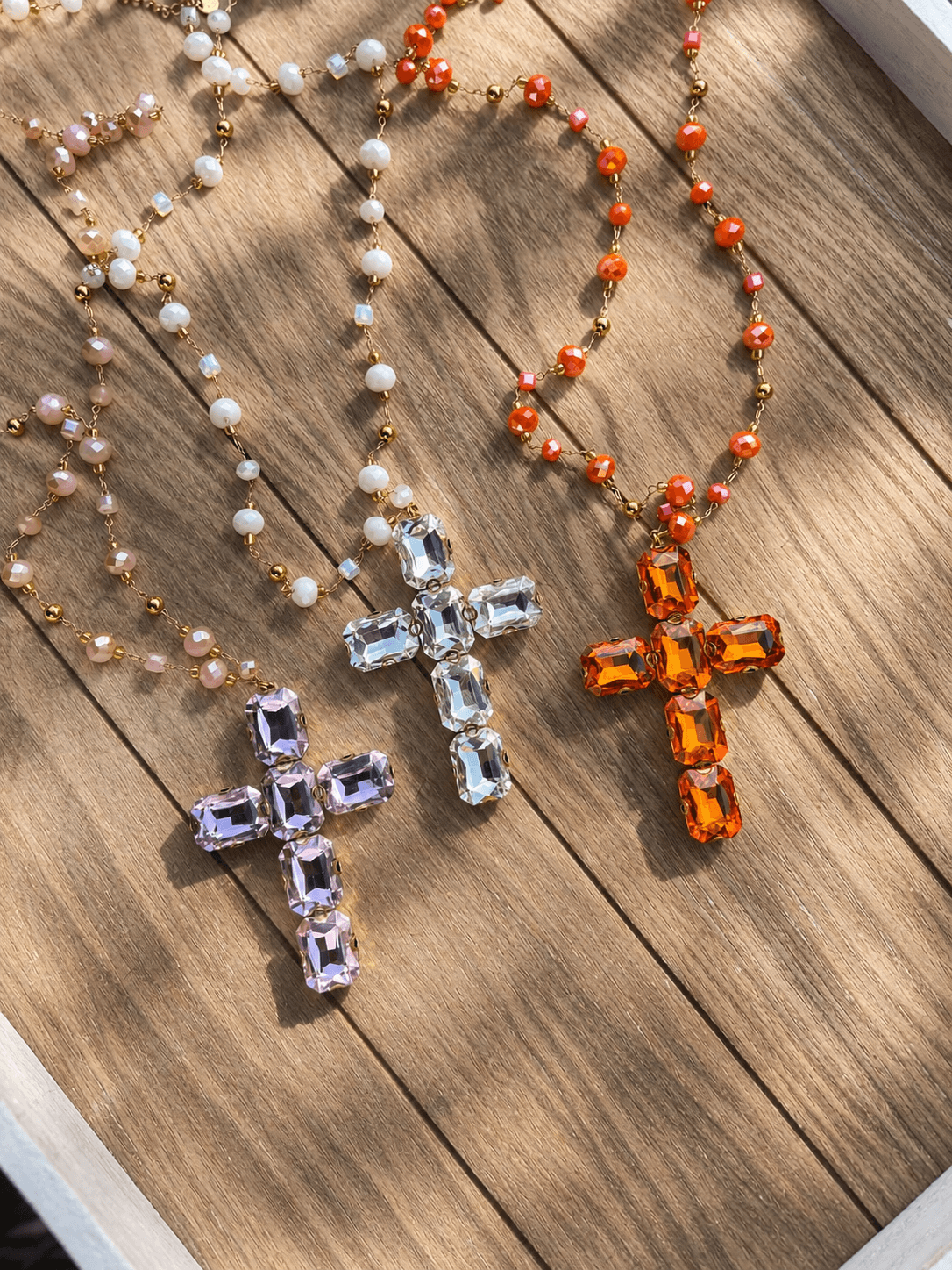 Drei elegante Kreuzketten mit filigraner Edelstahlkette und Perlen in Rosé, Crystal Light und Amber Glow auf einer Holzoberfläche.