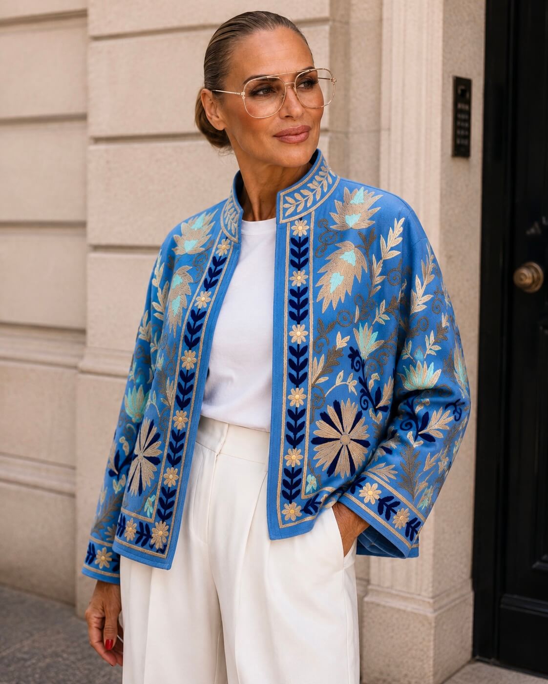 : Frau trägt eine blaue bestickte Couture Jacke von CASAPARI mit weißem Shirt und heller Hose vor einer eleganten Steinfassade.