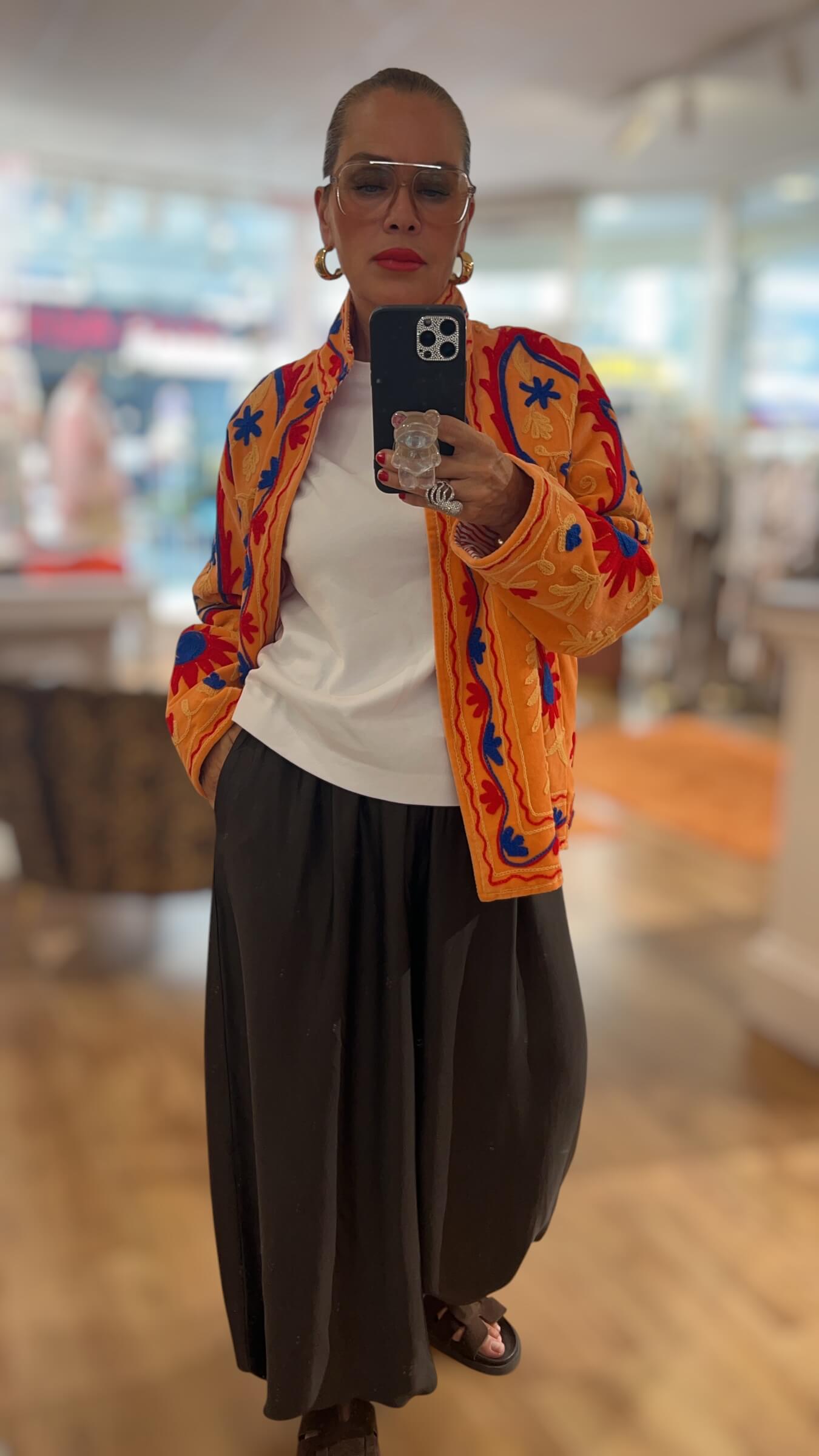 Frau trägt eine bestickte Couture-Jacke in warmen Gold-, Orange-, Rot- und Blautönen mit Gürtel kombiniert zu einer weiten schwarzen Hose.