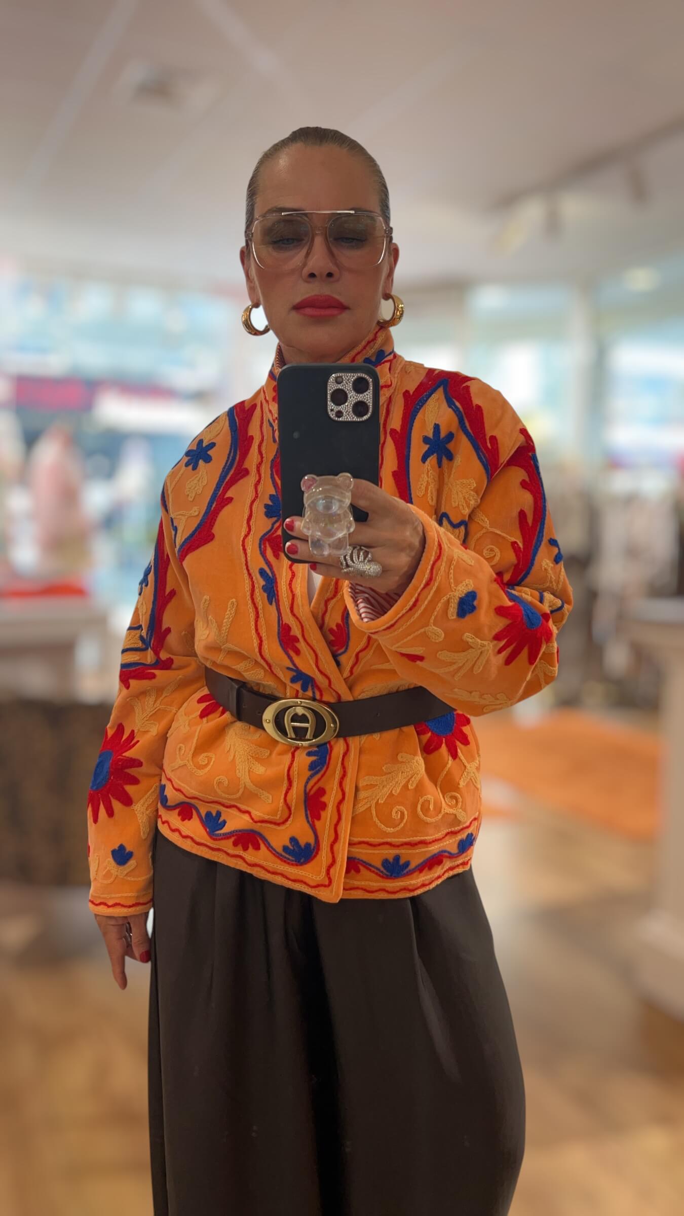 Frau trägt eine bestickte Couture-Jacke in warmen Gold-, Orange-, Rot- und Blautönen mit Gürtel kombiniert zu einer weiten schwarzen Hose.