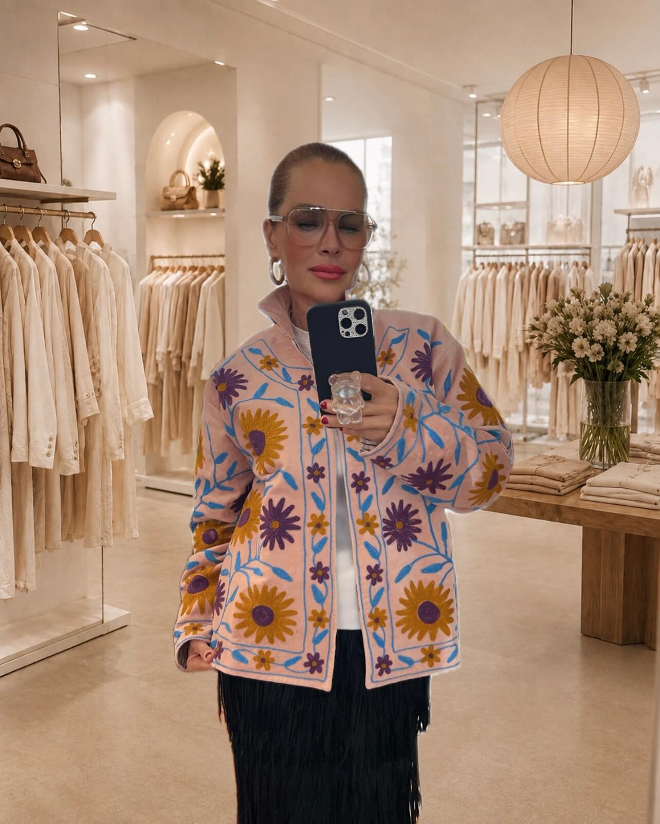 Frau fotografiert sich in einem Spiegel einer hellen Boutique. Sie trägt eine hellrosa, floral bestickte Embroidered Couture Jacke über einem weißen Oberteil und einem schwarzen Fransenrock. Im Hintergrund sind helle Kleiderstangen, ein Holztisch mit gefalteter Kleidung und ein Blumenstrauß zu sehen.