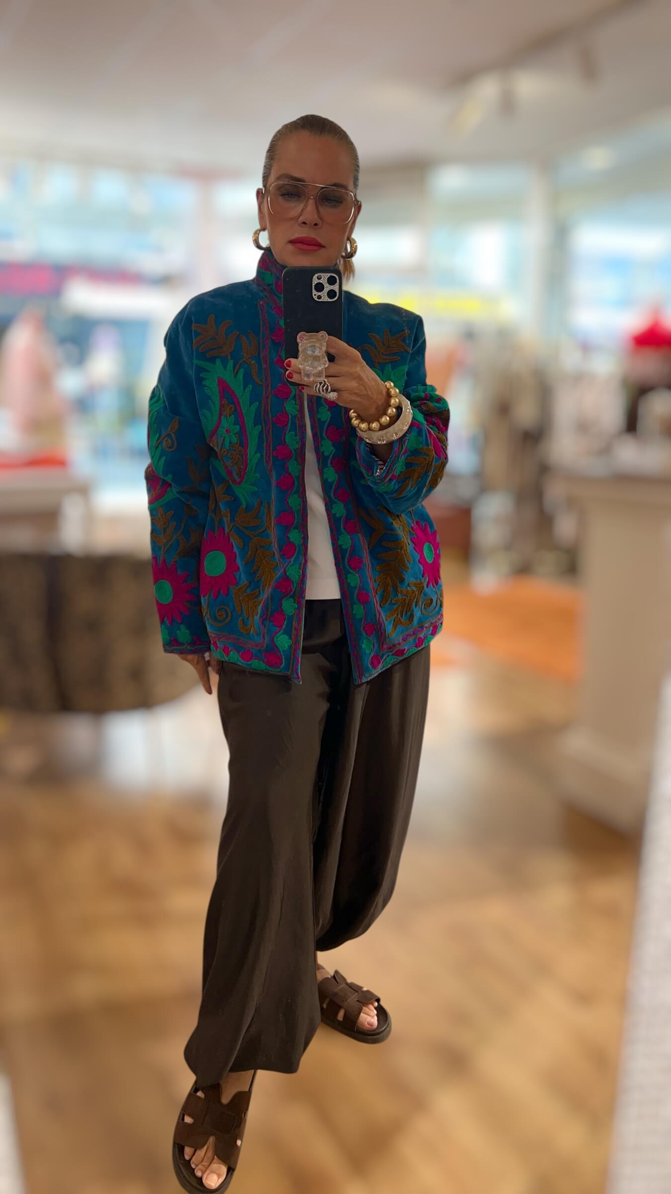 Frau trägt die Embroidered Couture Jacke Saphir Fuchsia in Petrolblau mit aufwendigen Stickereien in Fuchsia, Türkis und Olivgrün, kombiniert mit weißem Shirt und weiter schwarzer Hose.