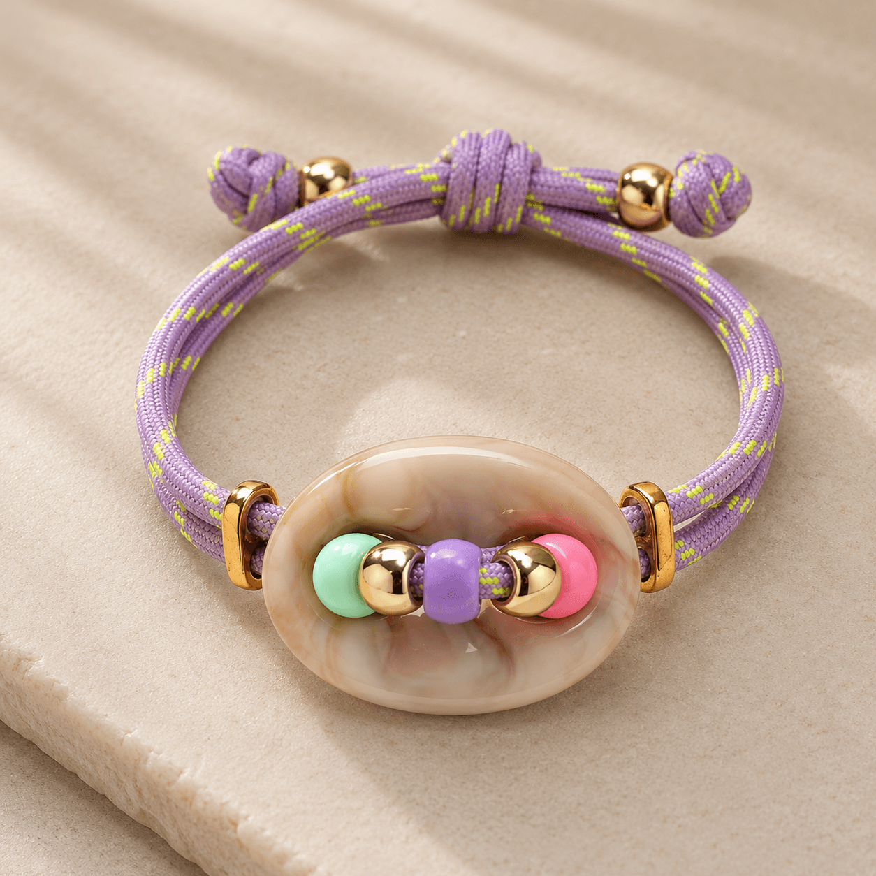 Verstellbares Armband mit fliederfarbenem Baumwollband, cremefarbener Resina-Perle sowie bunten Resina- und goldfarbenen Edelstahl-Elementen.