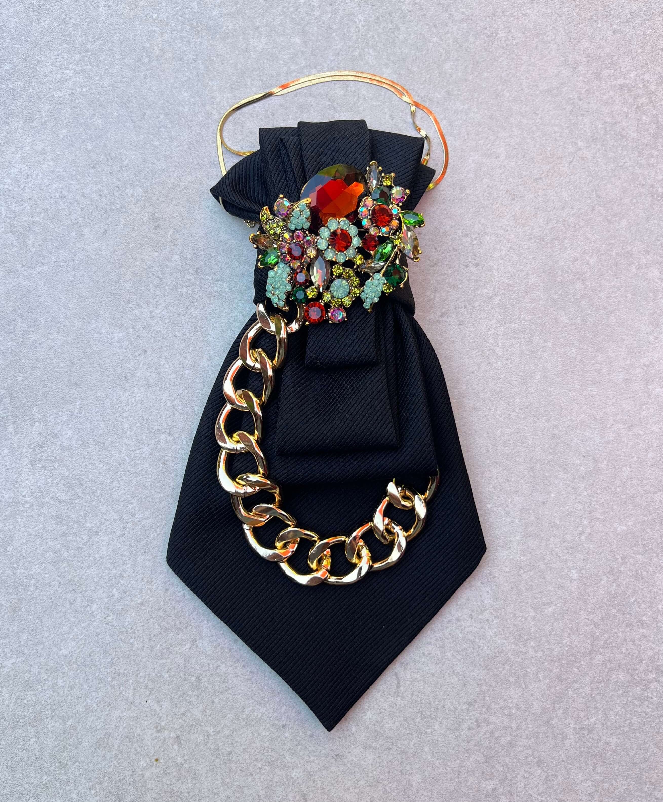 Breite schwarze Vintage-Krawatte mit goldfarbener Kette und farbiger Schmuckbrosche auf heller Steinplatte.