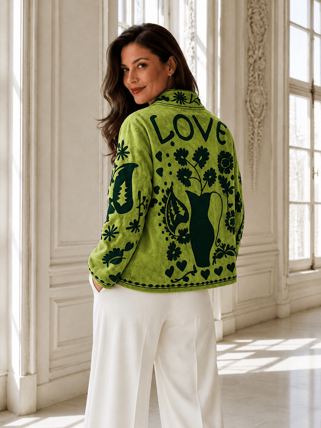 Frau in heller Hose trägt eine grüne Jacke mit großflächigem floralem Muster und Schriftzug auf dem Rücken in einem hellen eleganten Innenraum, Rückenansicht.