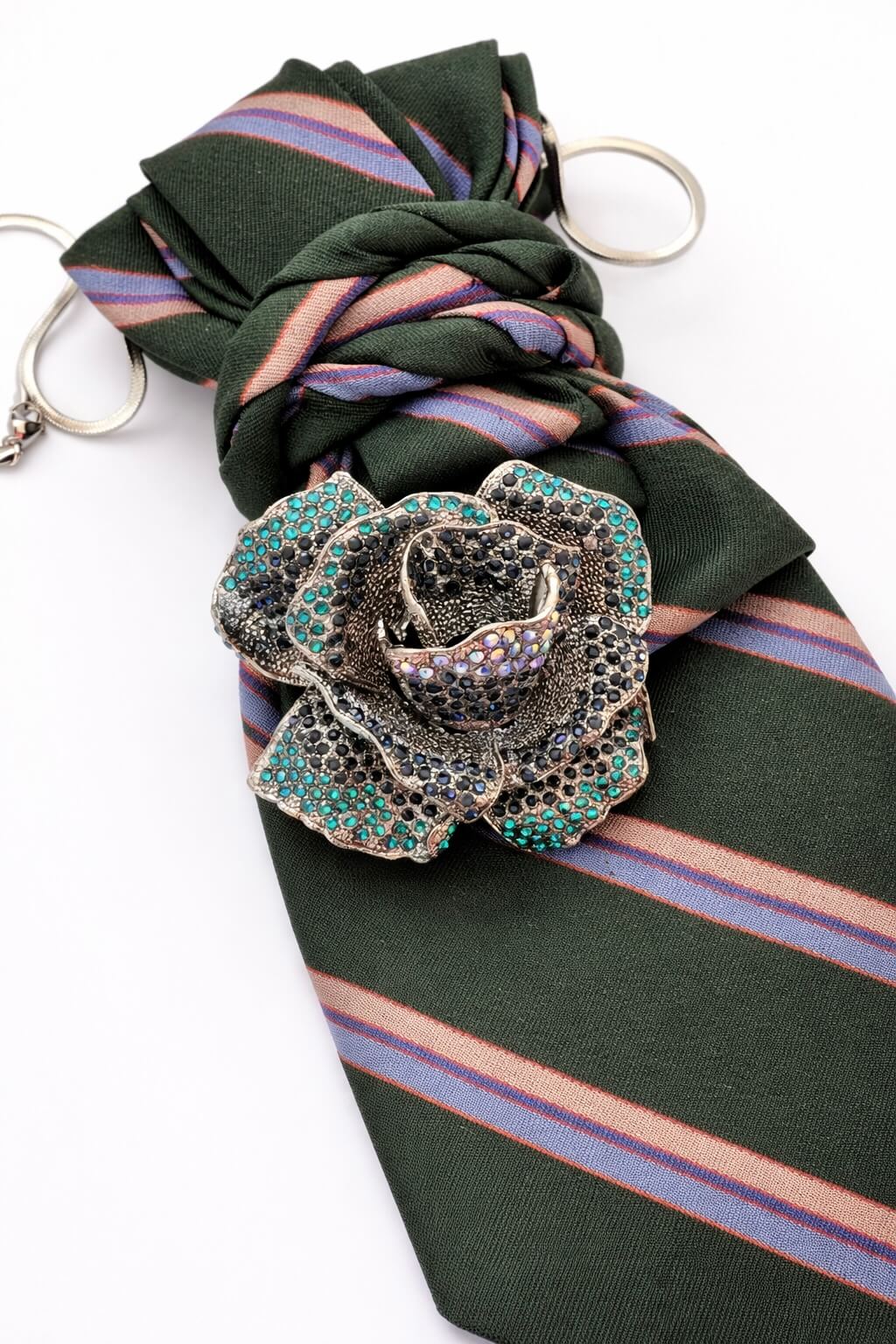 Grüne Krawatte mit diagonalen Streifen, silberfarbener Kette und großer Rosenbrosche mit blauen und dunklen Steinen, dekorativ gelegt auf hellem Hintergrund