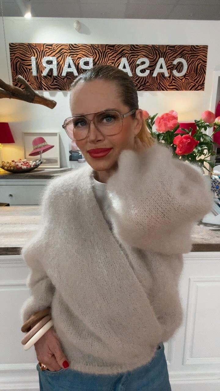Frau trägt einen hellen, weichen Strick-Cardigan in Wickeloptik mit Jeans und Schmuck, stehend in einer Boutique.