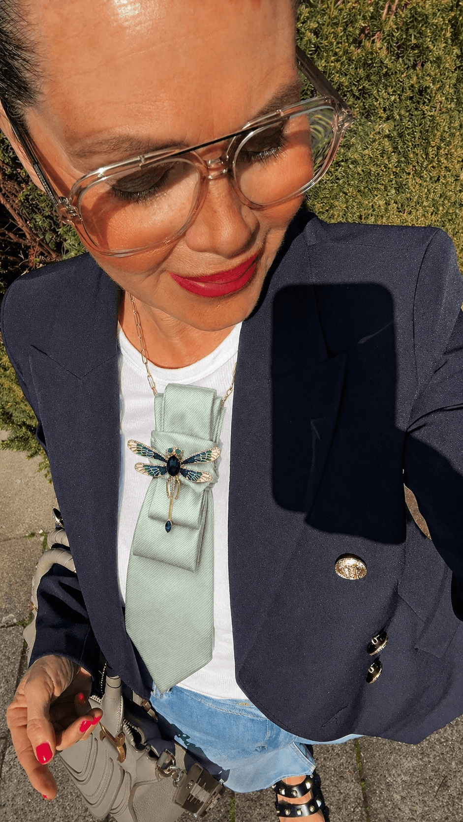 Frau macht ein Selfie im Freien, trägt eine große transparente Brille, dunkelblauen Blazer über weißem Oberteil sowie eine mintfarbene Krawatte mit Libellenbrosche, dazu rote Nägel und eine Handtasche.