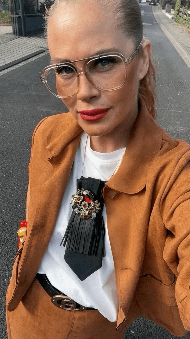 Frau steht auf einer Straße. Sie hat Cognac, farbenfroh Rock und Cognac, farbenfroh Jacke an weißes T-Shirt und trägt eine auffällige Krawatte mit Brosche und schaut in die Kamera.