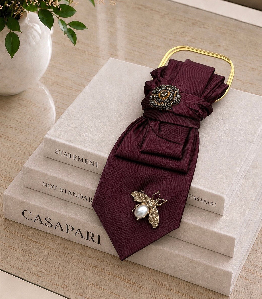 Bordeauxfarbene Statement-Krawatte von Casapari mit goldfarbener Kette, floraler Brosche und Bienenbrosche auf Holzboden.