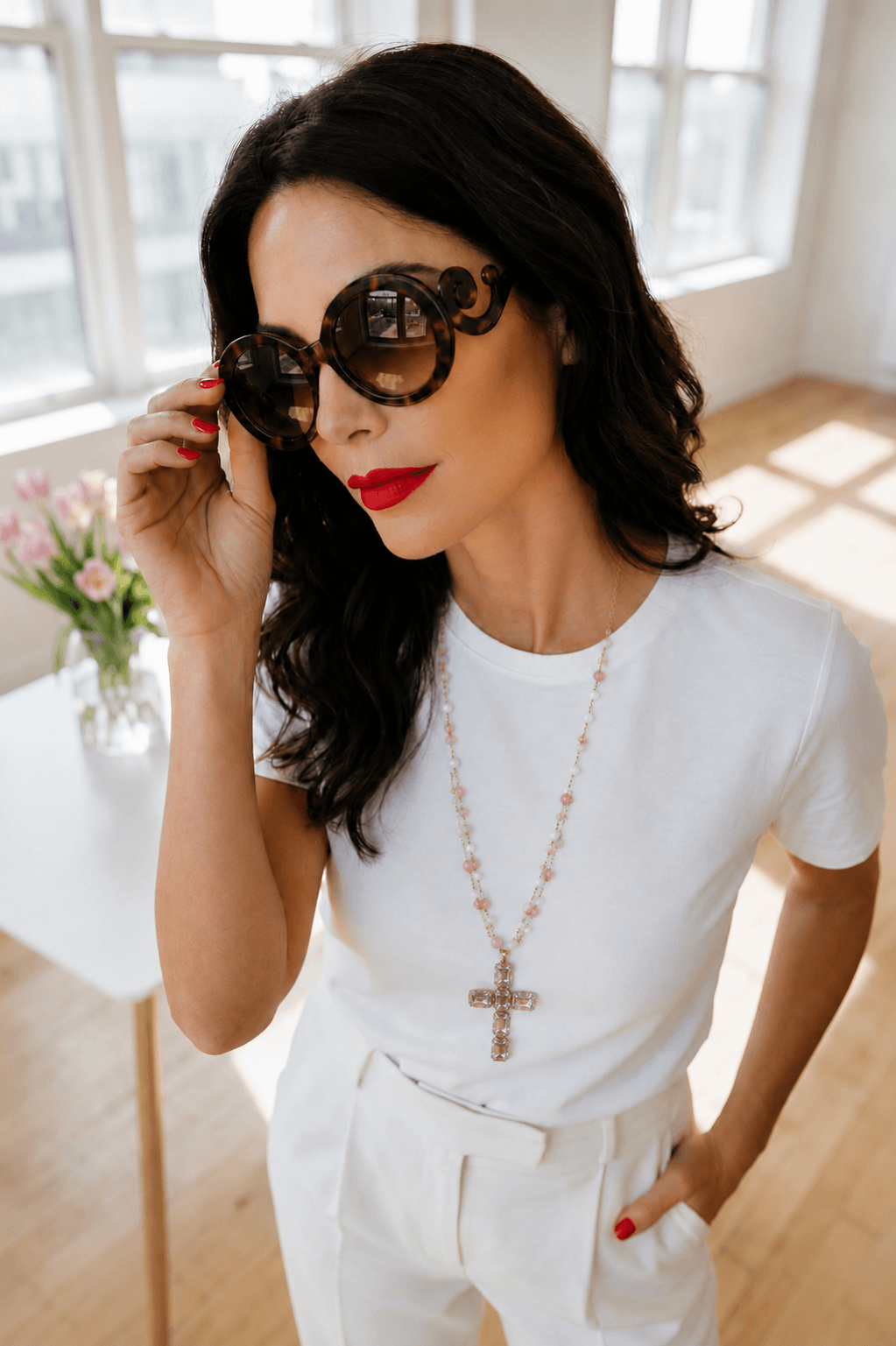 Frau steht in einem Kellerraum sie trägt eine weiße Hose weißes T-Shirt unten eine rosafarbene lange Kette mit Kreuz und sie trägt eine Sonnenbrille