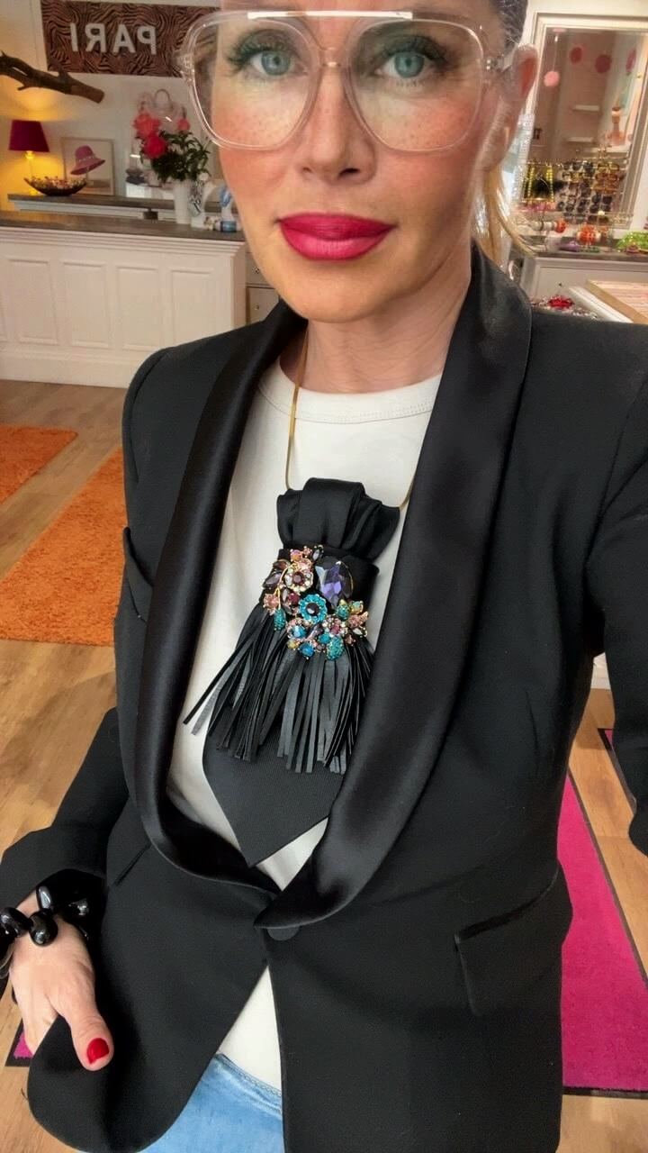 Frau trägt eine schwarze Vintage-Krawatte mit dekorativer Brosche und Fransen-Detail zu schwarzem Blazer in einer Boutique.
